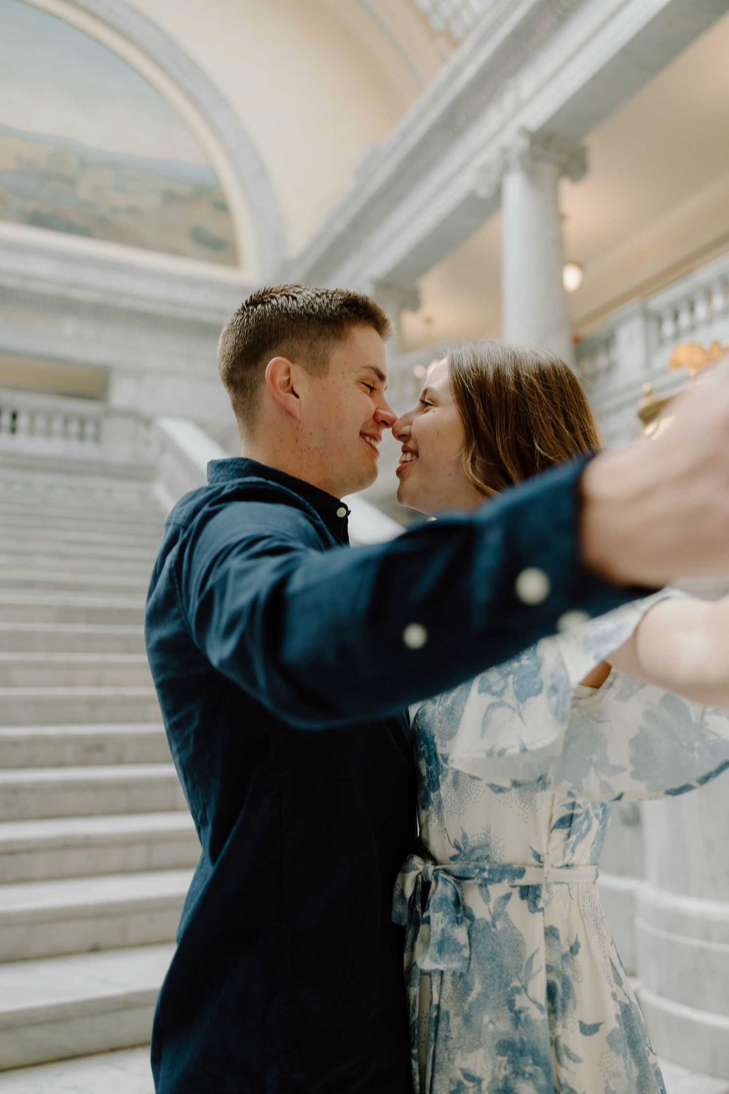 Couple sharing a romantic nose-to-nose moment on grand marble staircase in elegant building.