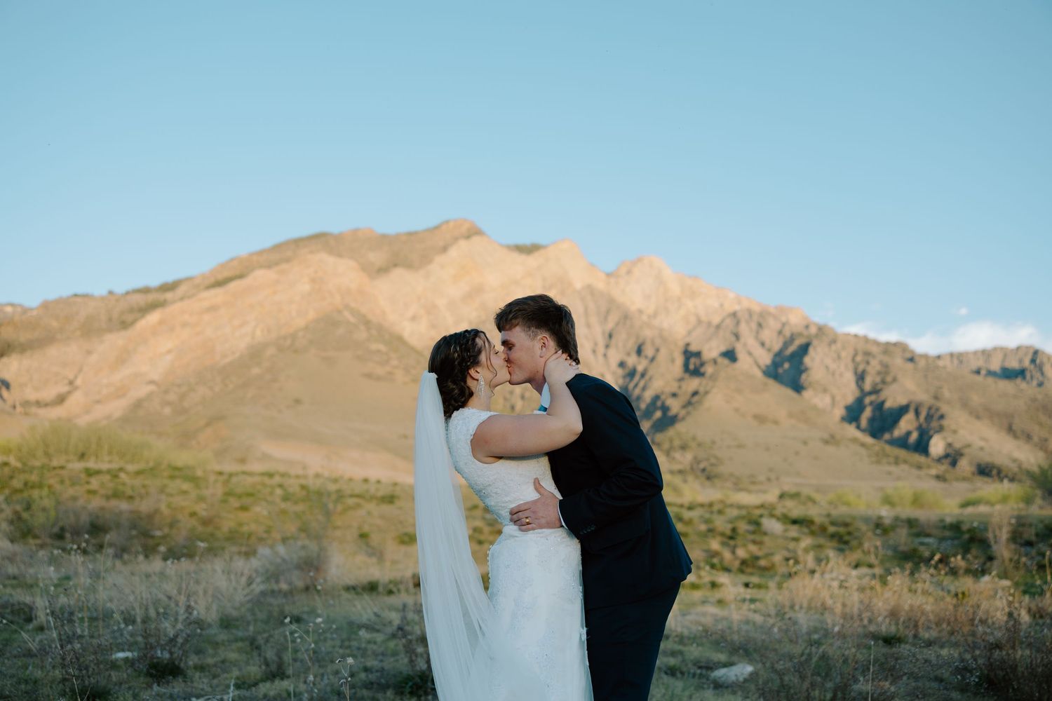 Wedding couple kissing with stunning mountain backdrop under clear blue sky in an outdoor ceremony.