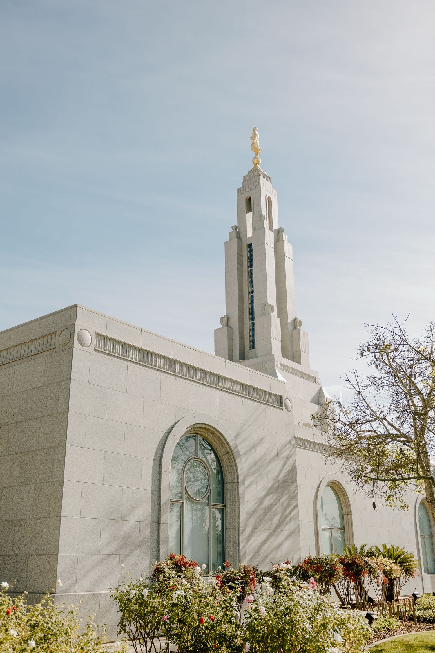 White LDS Mormon temple with gold angel Moroni spire against blue sky, surrounded by lush green gardens and flowers.