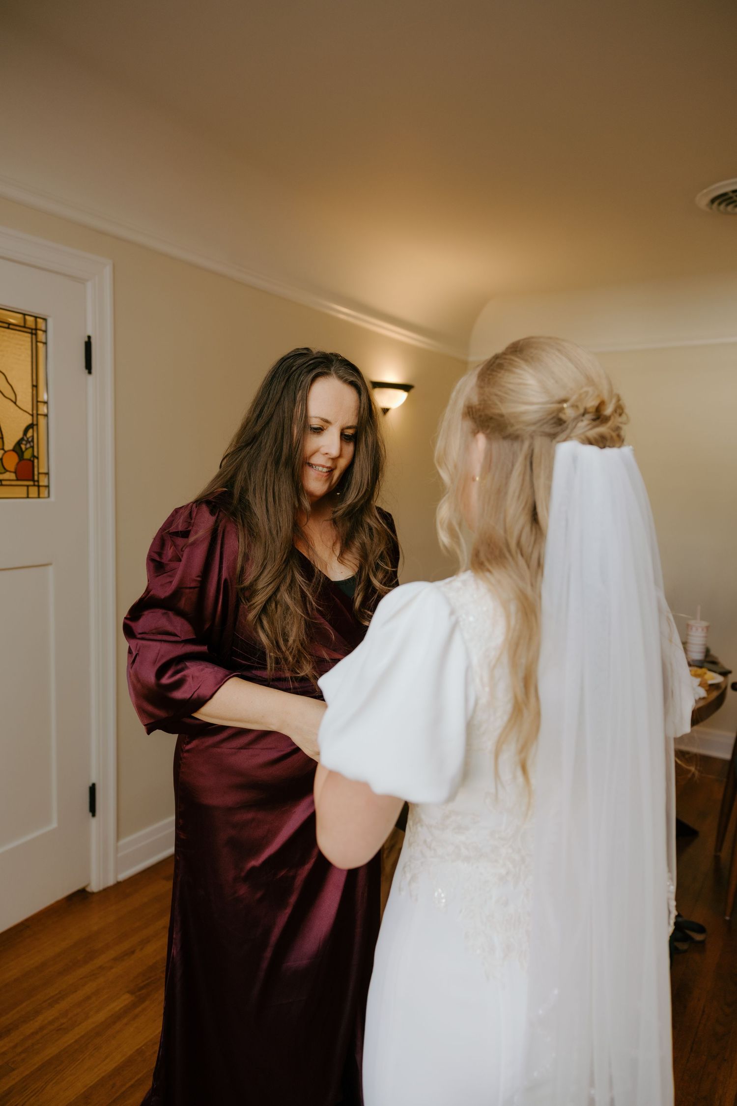 Bridesmaid in burgundy satin dress helps blonde bride in white lace gown prepare indoors before wedding.