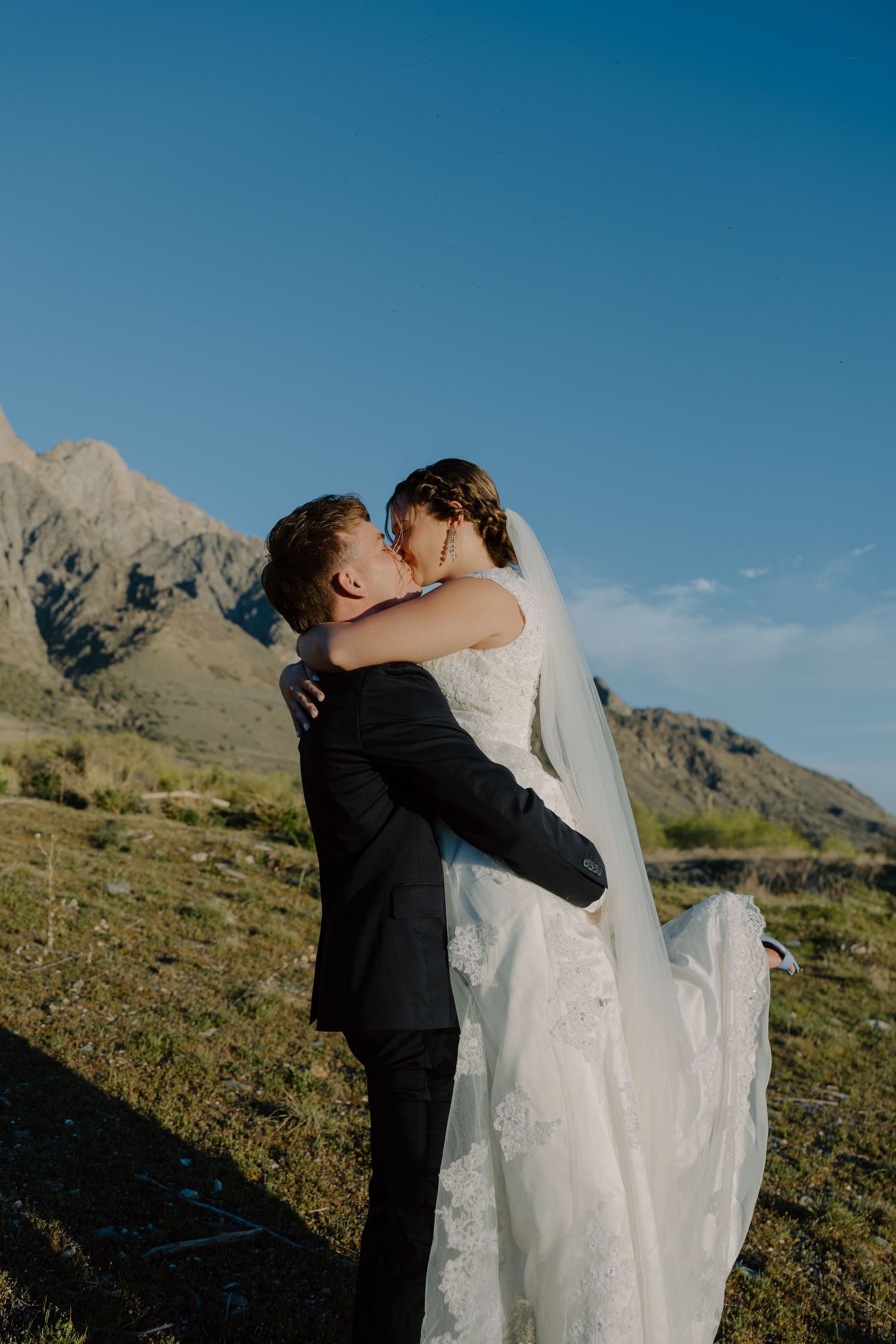 Groom lifting bride in white gown and veil, sharing a kiss with mountain landscape and blue sky backdrop.