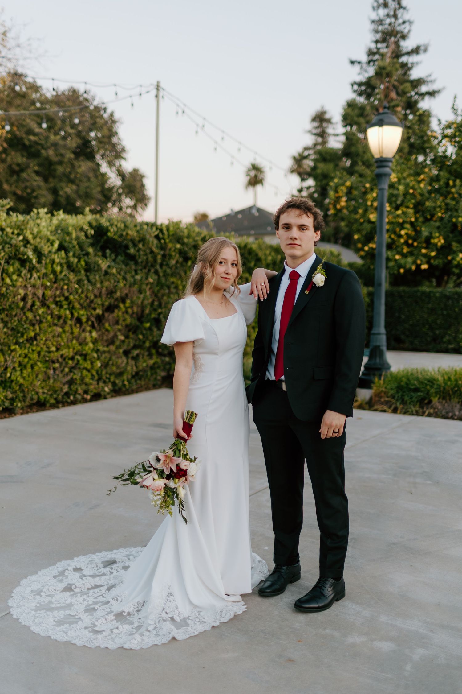Bride in white lace gown holding pink bouquet stands with groom in black suit and red tie at outdoor wedding venue.
