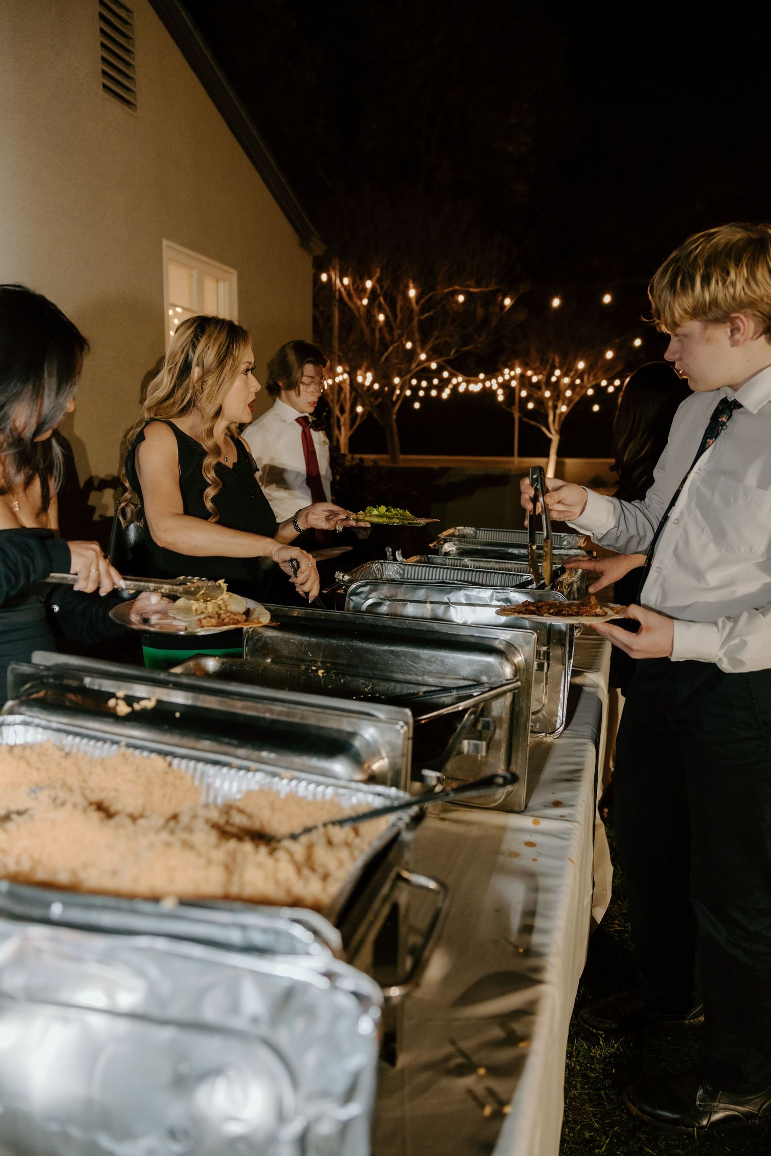 Wedding guests serve themselves at an outdoor buffet dinner under warm string lights at a nighttime reception.