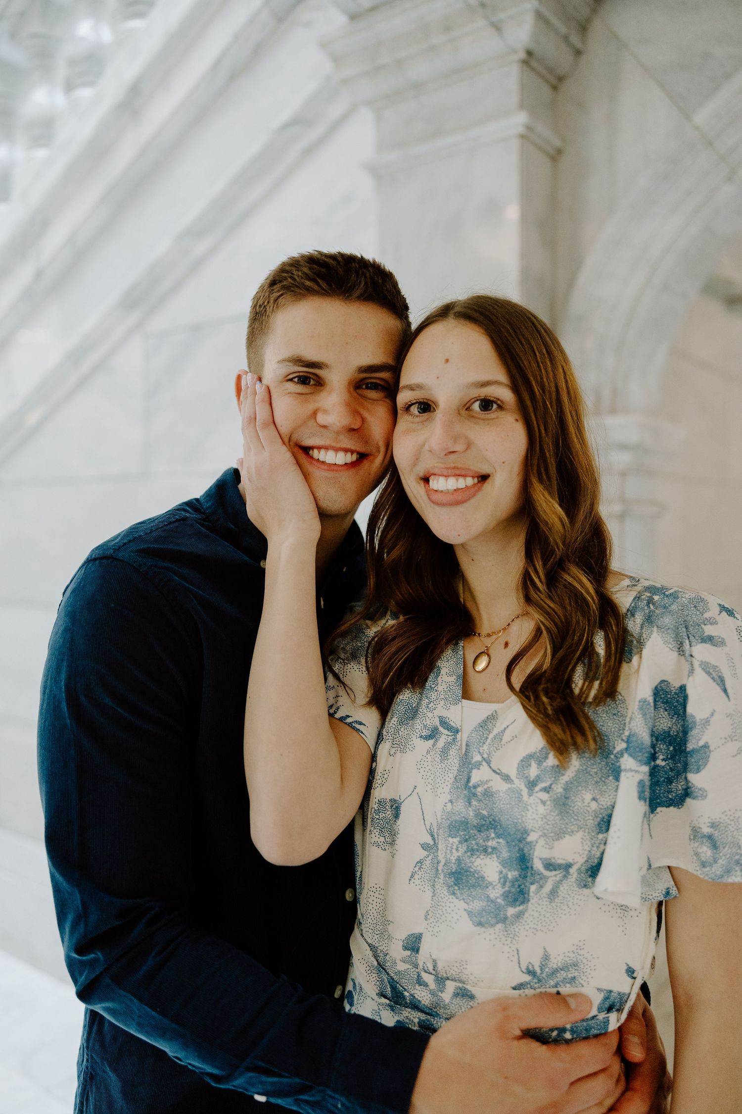 A happy couple poses cheek-to-cheek indoors, woman in blue floral dress, man in navy shirt, smiling at camera.