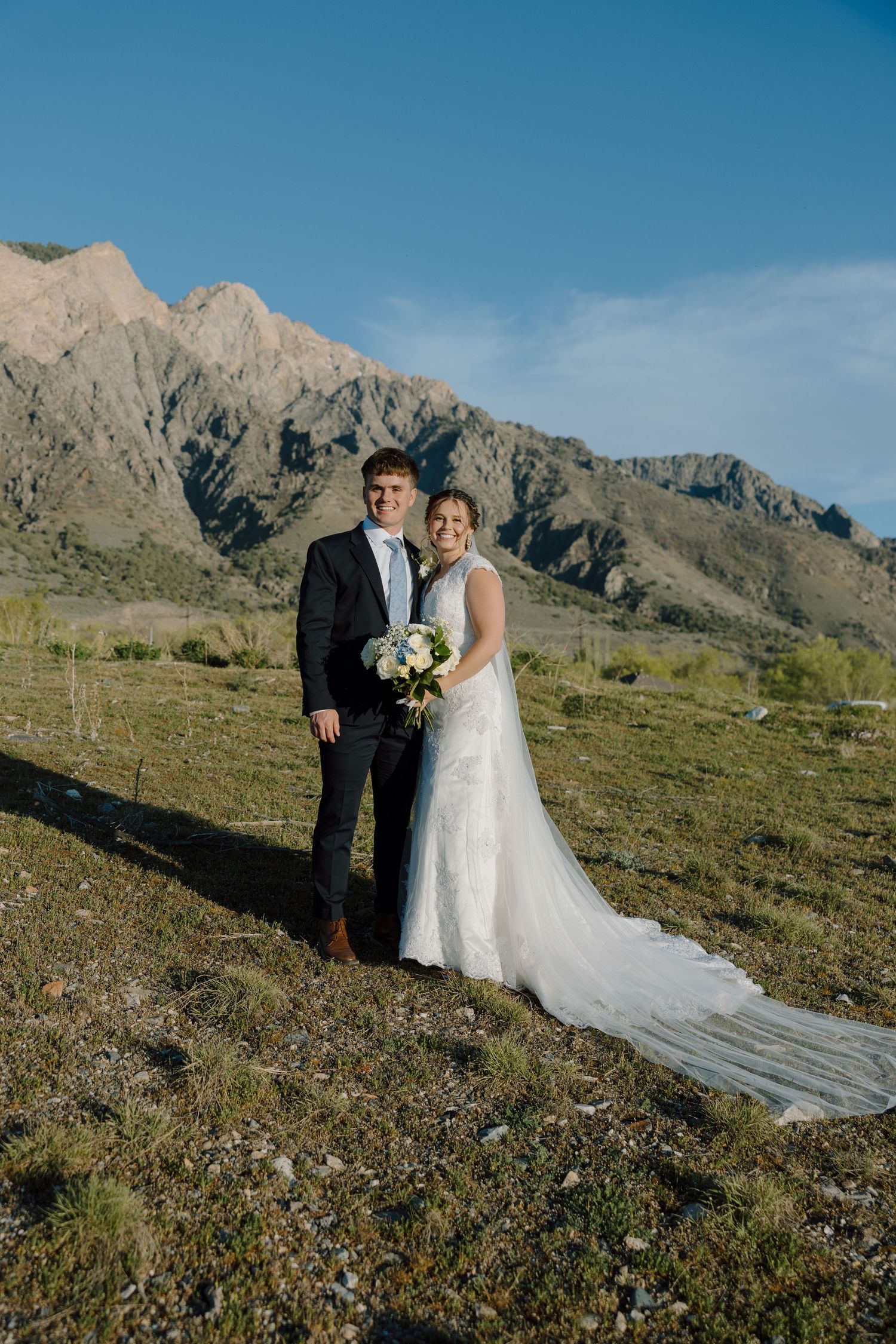 Wedding couple poses together in lace gown and dark suit with dramatic mountain scenery behind them.