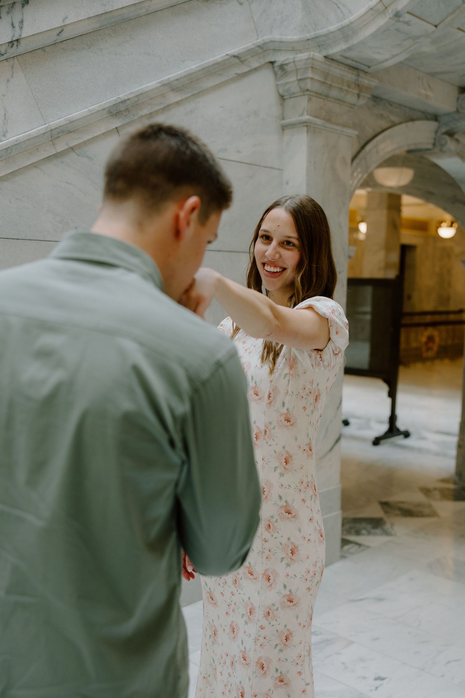 Smiling woman in floral dress holds mans hand while dancing in a grand marble hallway.
