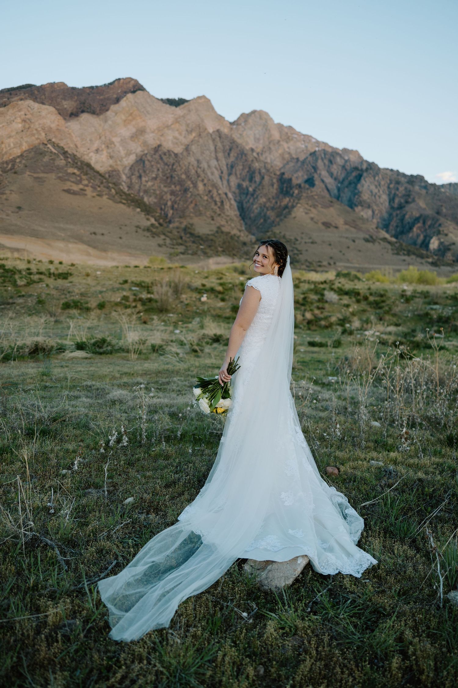 Bride in white gown with veil holding bouquet, posing in mountain meadow landscape at golden hour.