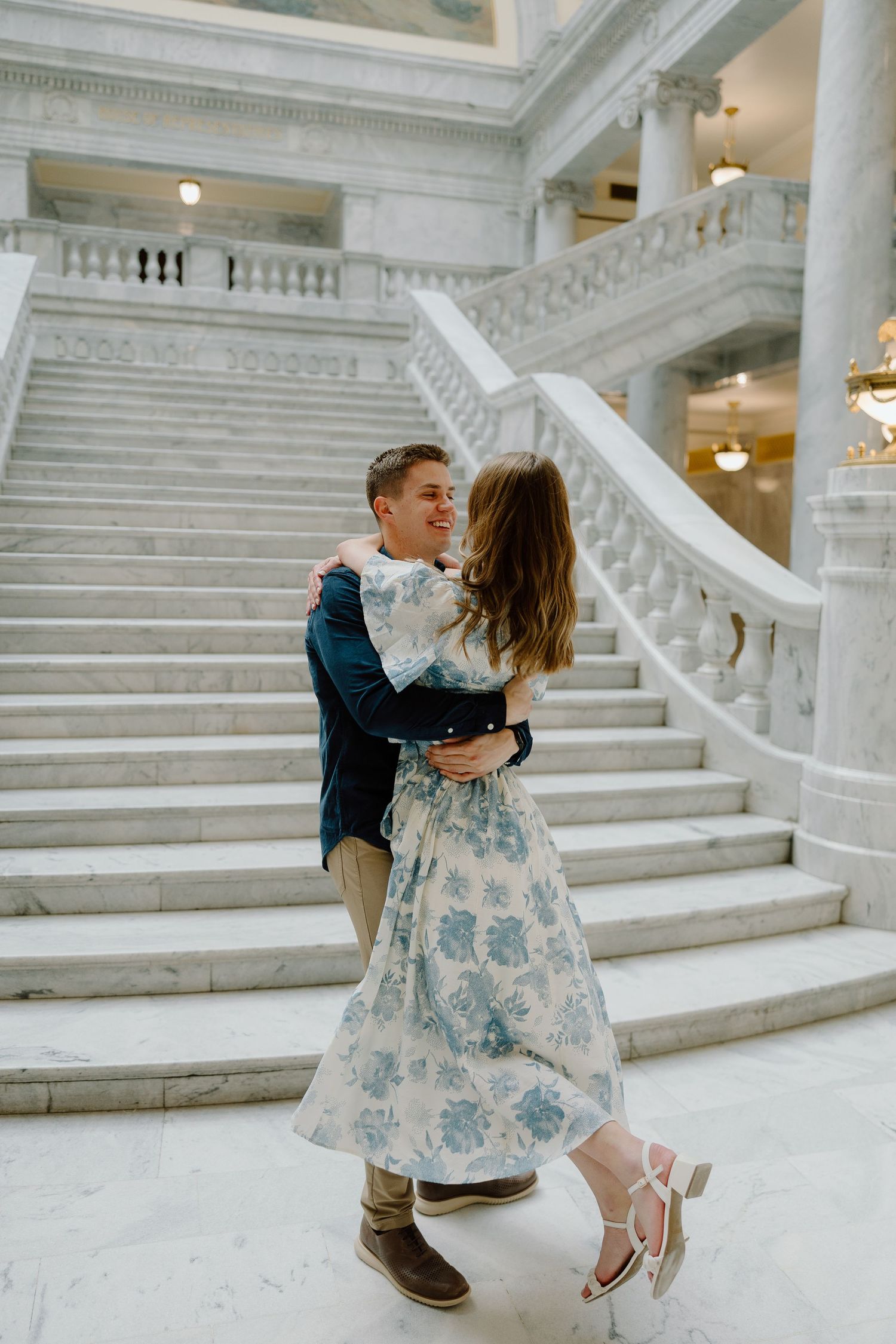 Couple embracing and dancing on marble stairs in an elegant building, woman in floral dress, man in navy blazer.