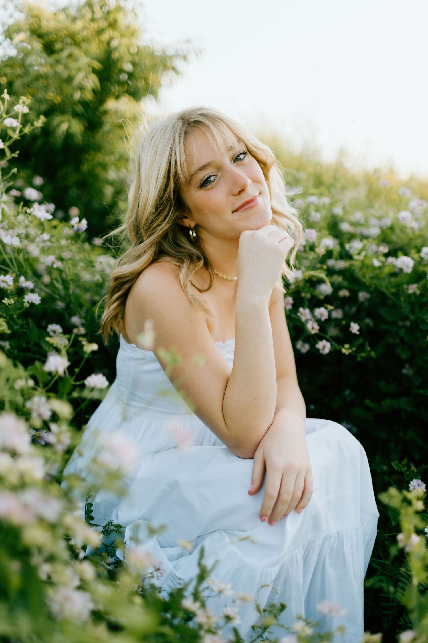 Young blonde woman in white dress sitting among pink wildflowers, smiling softly in golden sunlight.