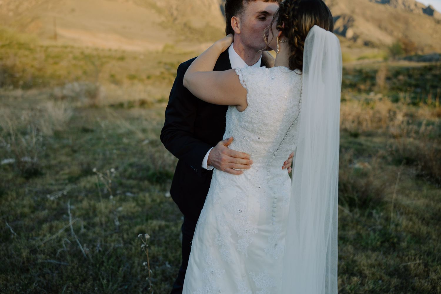 Bride and groom sharing a romantic kiss in a golden field with rolling hills at sunset on their wedding day.