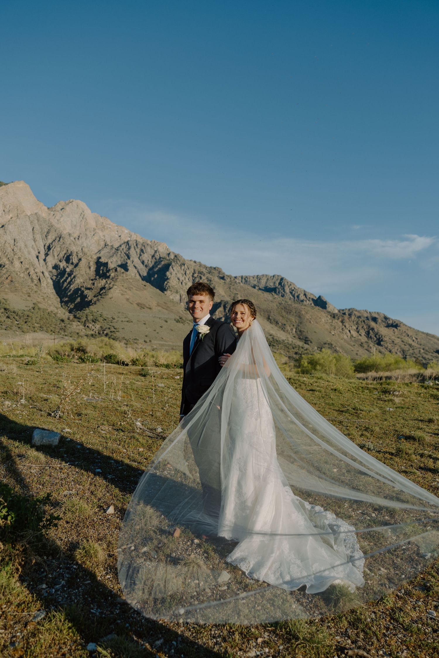 Smiling newlyweds posing with bride's veil billowing in the breeze before mountain backdrop.