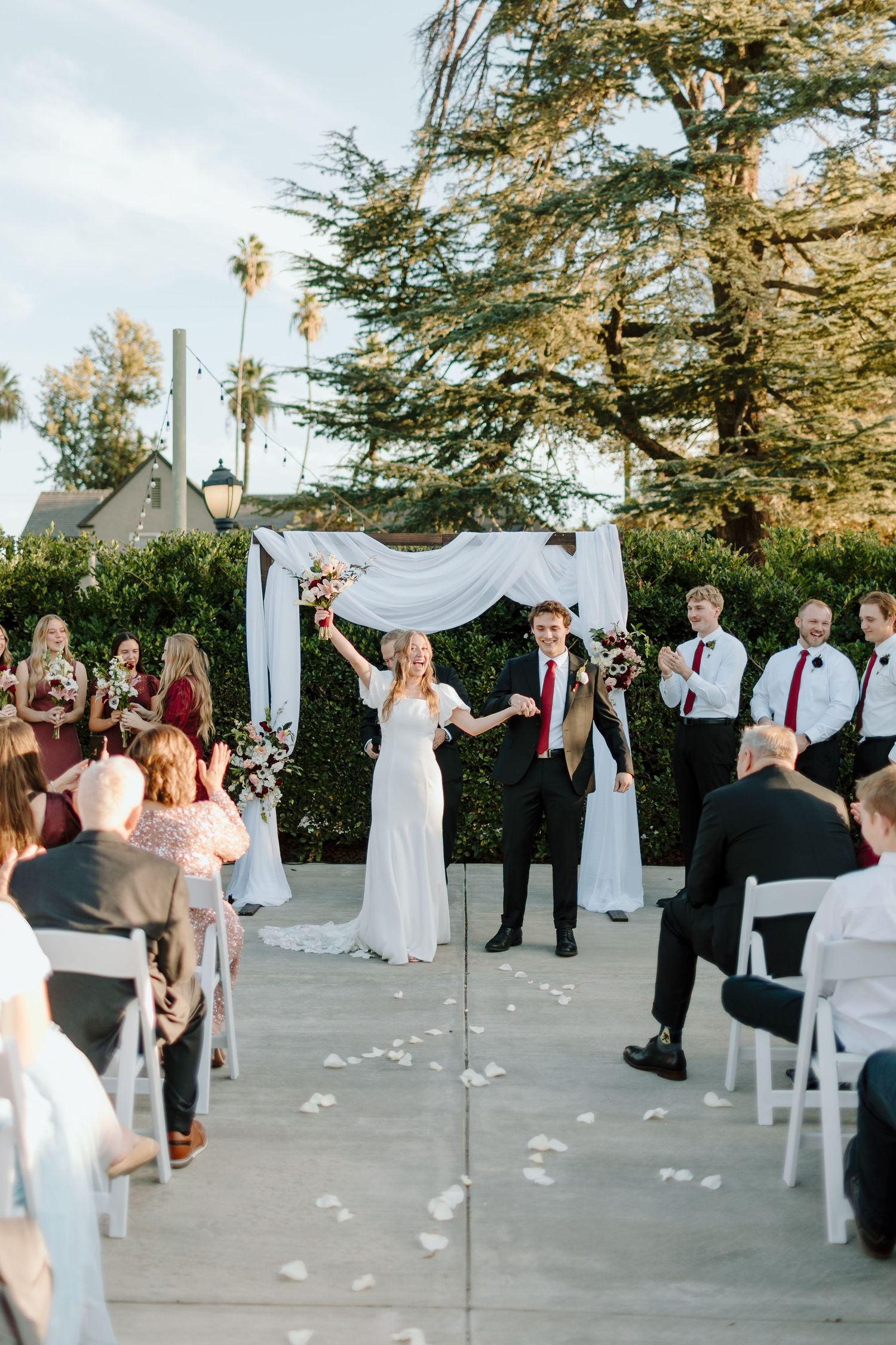 Bride and groom walk down the aisle at an outdoor wedding ceremony with white floral arch and seated guests.