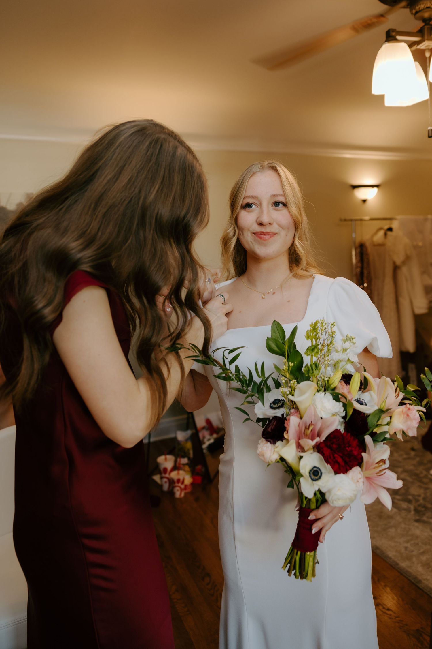 Smiling bride in white square-neck gown holds pink and burgundy floral bouquet while chatting with bridesmaid.