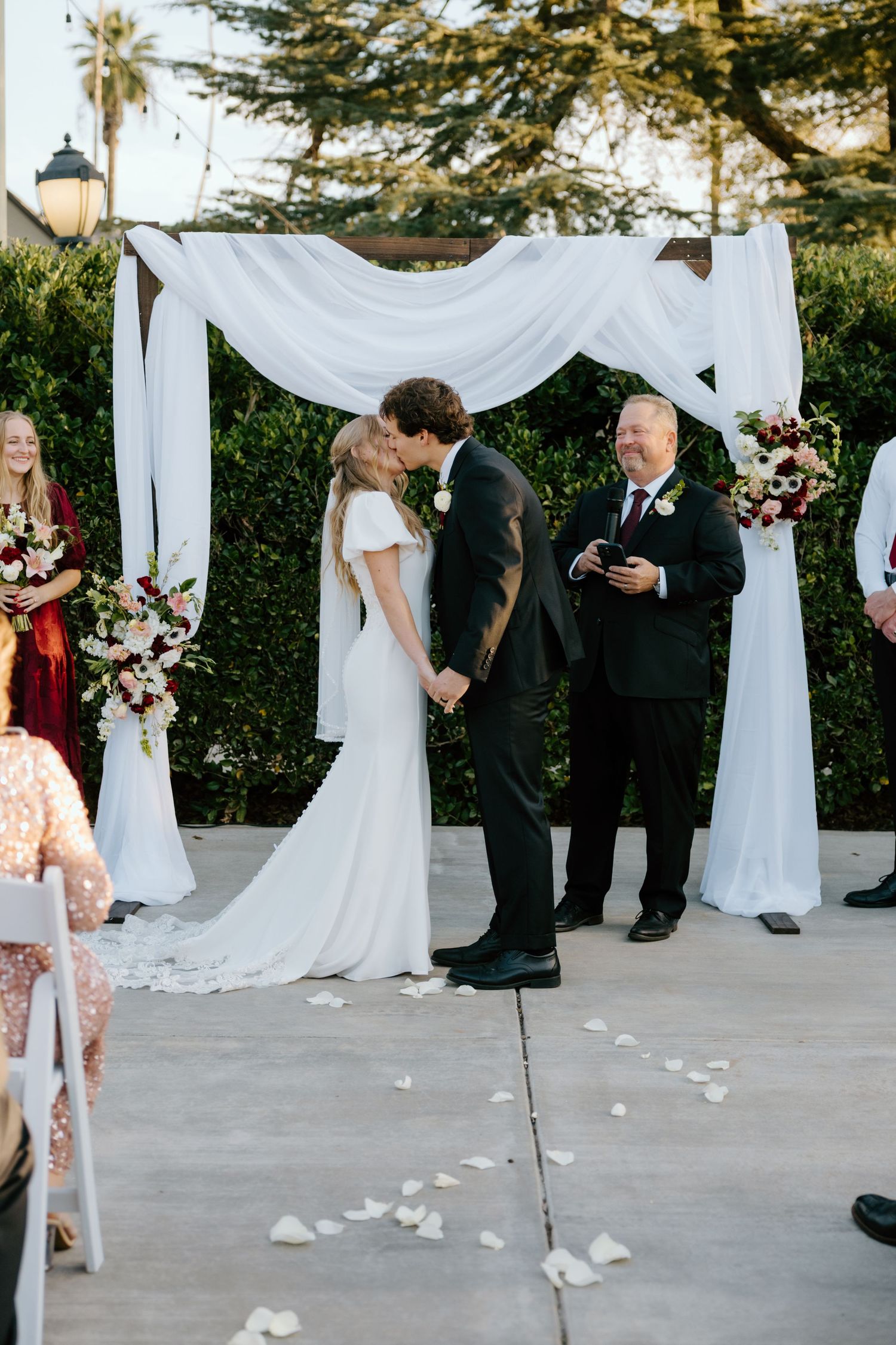 Bride and groom share first kiss under white draped arch with rose petals on ground at outdoor ceremony.