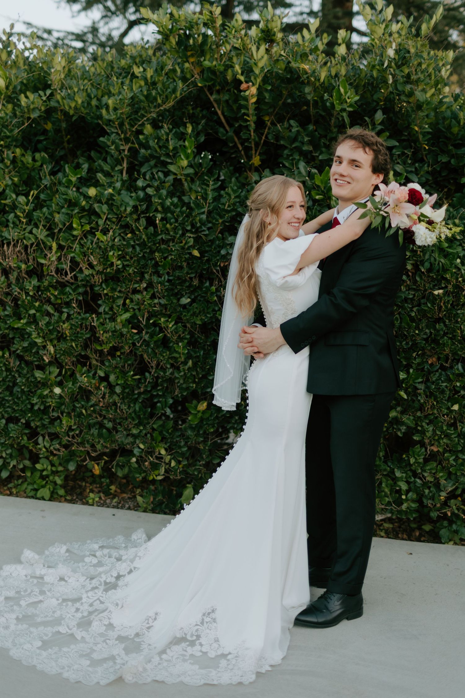 Bride and groom embracing outdoors, bride in white lace gown with veil holding bouquet, groom in dark suit, green hedge backdrop.