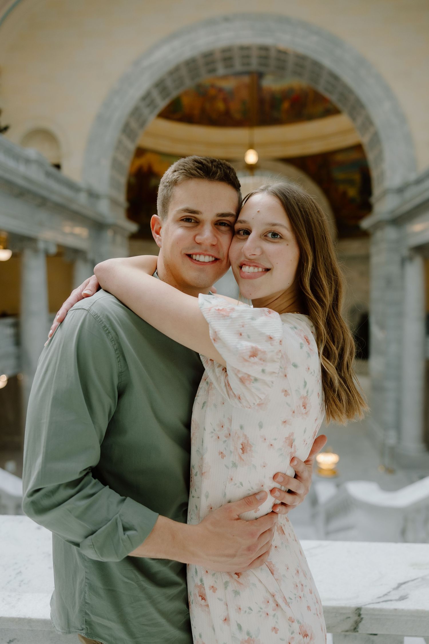 A smiling couple embraces in an elegant domed building, woman in floral dress hugging man in green shirt.