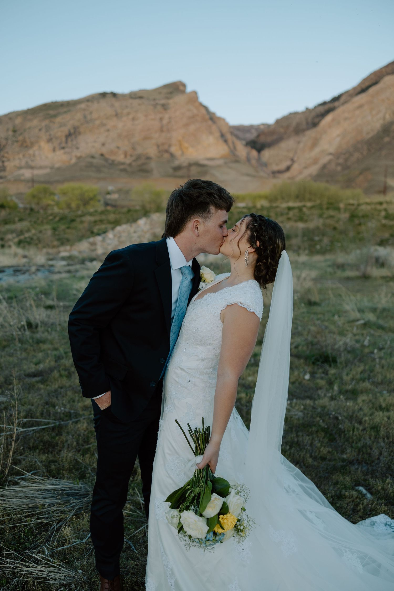 Bride and groom share a romantic kiss in a scenic desert landscape with golden mountain backdrop during outdoor wedding.