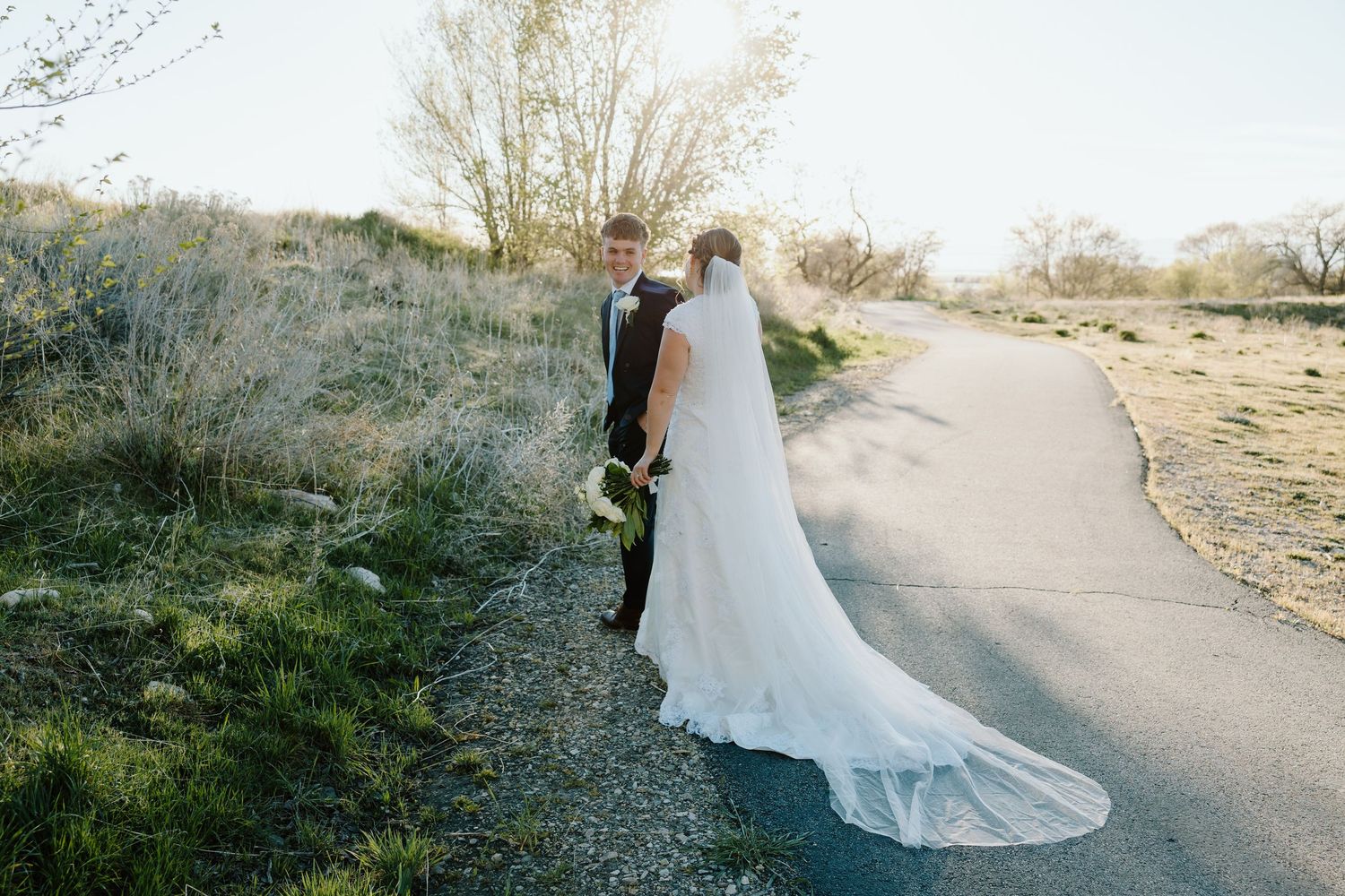 Bride in flowing white gown with long veil walking with groom on a scenic path.