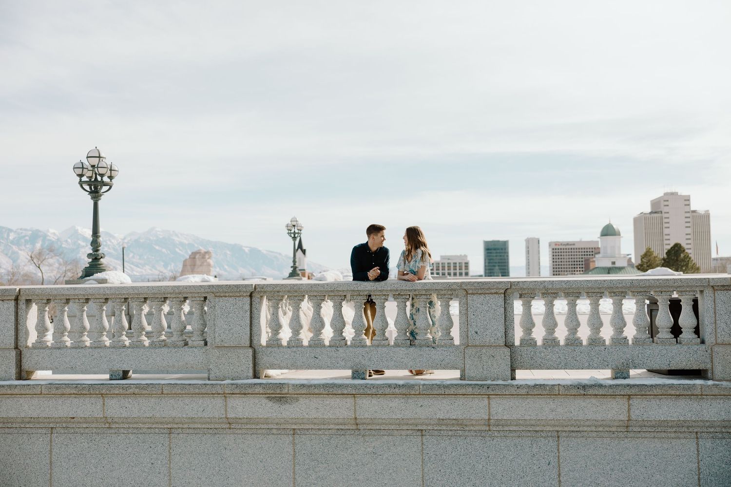 A couple stands together on a marble balcony overlooking a city skyline with snow-capped mountains behind.