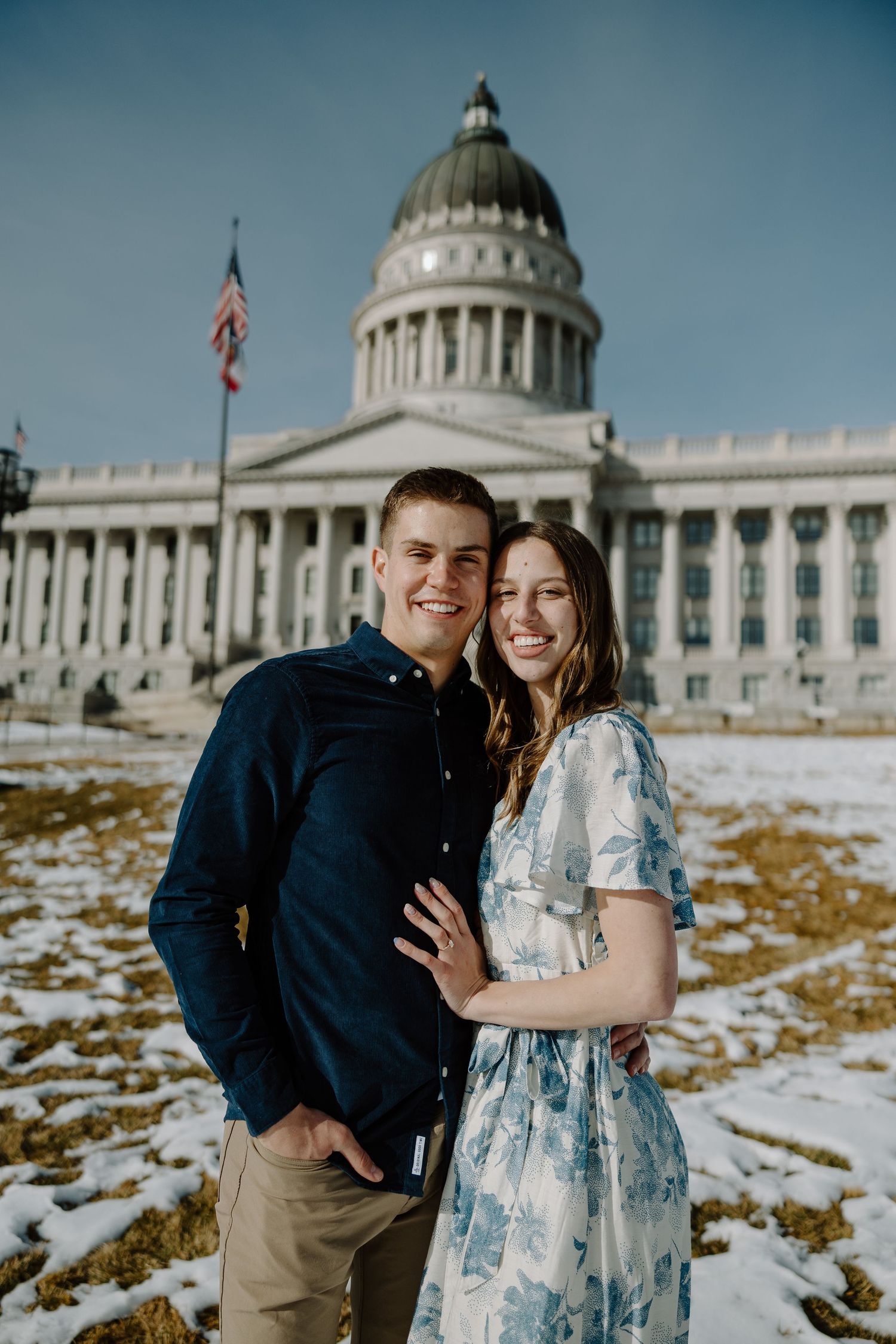 Smiling couple posing in front of Utah State Capitol building in winter with snow on ground.