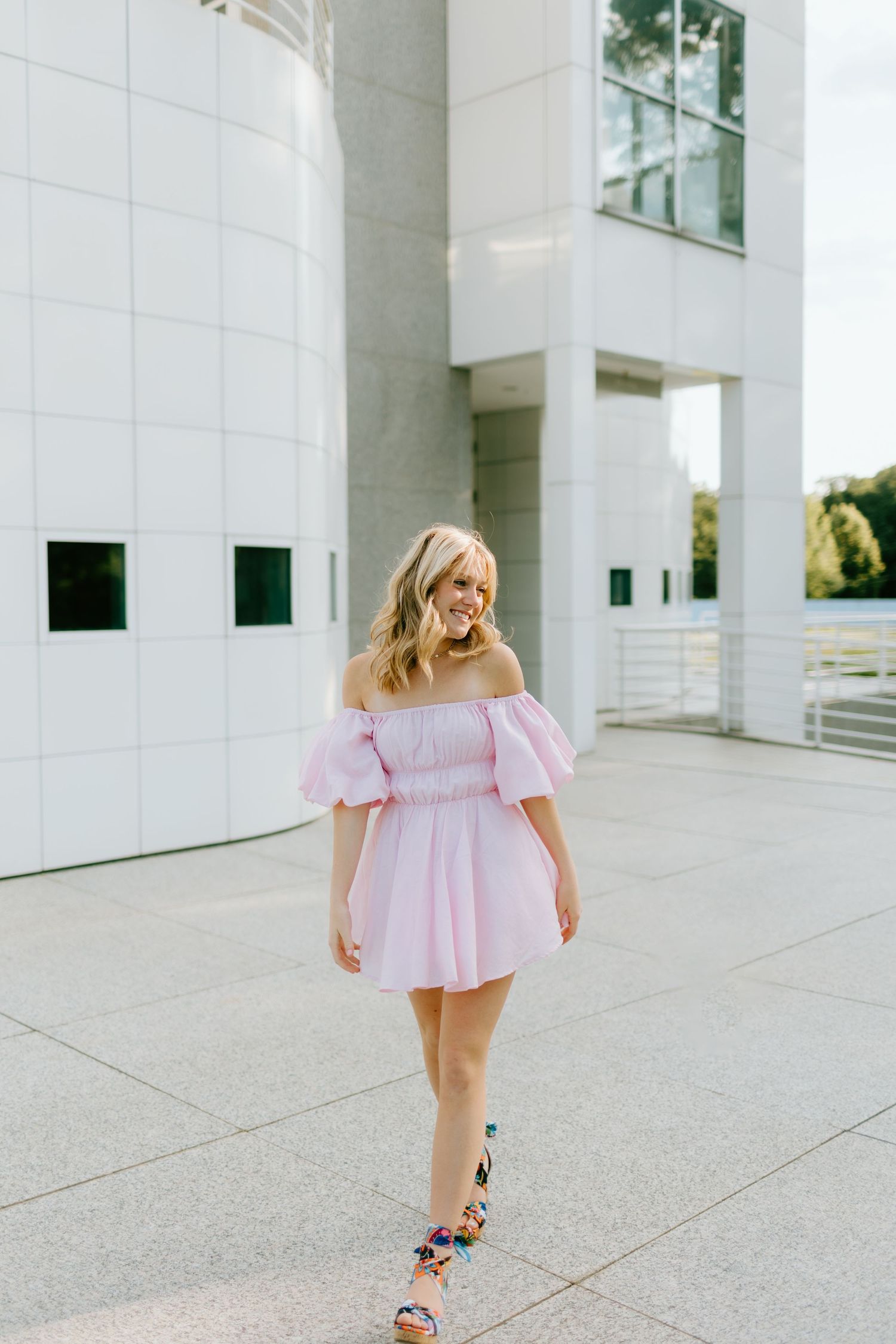 Smiling blonde woman walking in pink off-shoulder puff sleeve mini dress with colorful sandals near modern building.