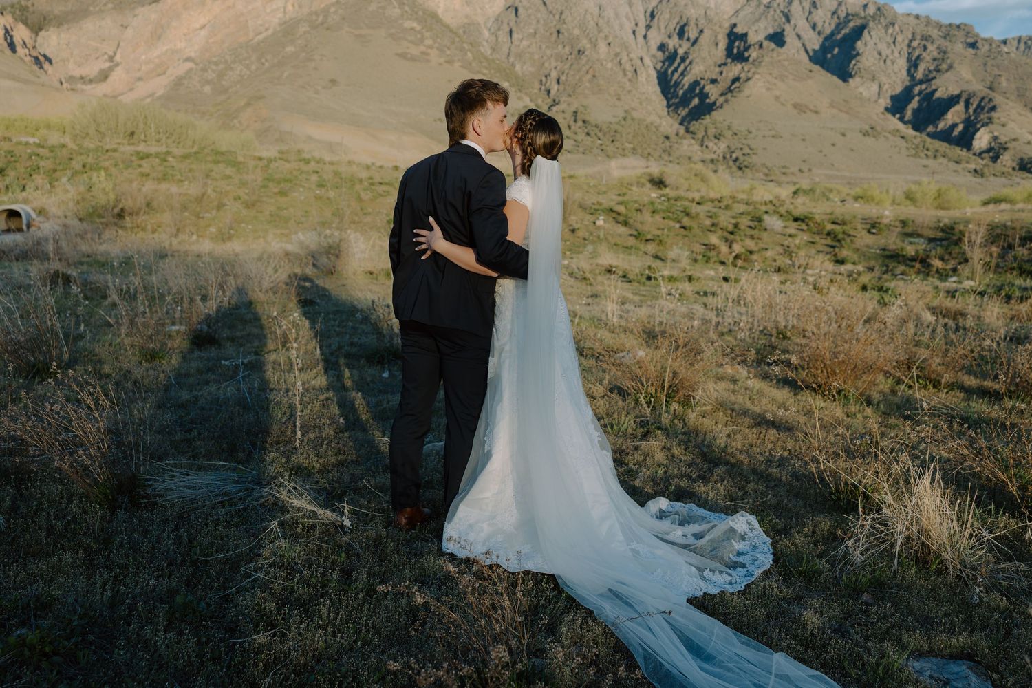 Bride and groom sharing a kiss from behind with long veil trailing across mountain landscape at sunset.
