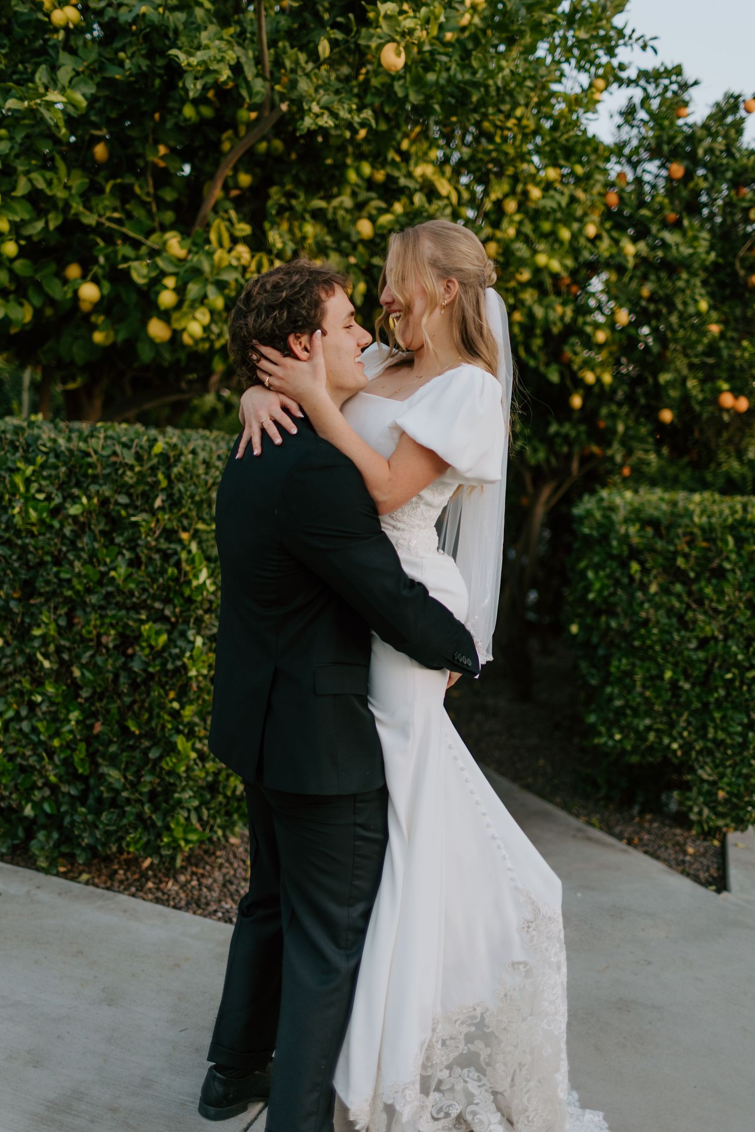 Bride and groom embracing tenderly in front of lush green hedges and citrus trees during outdoor wedding.