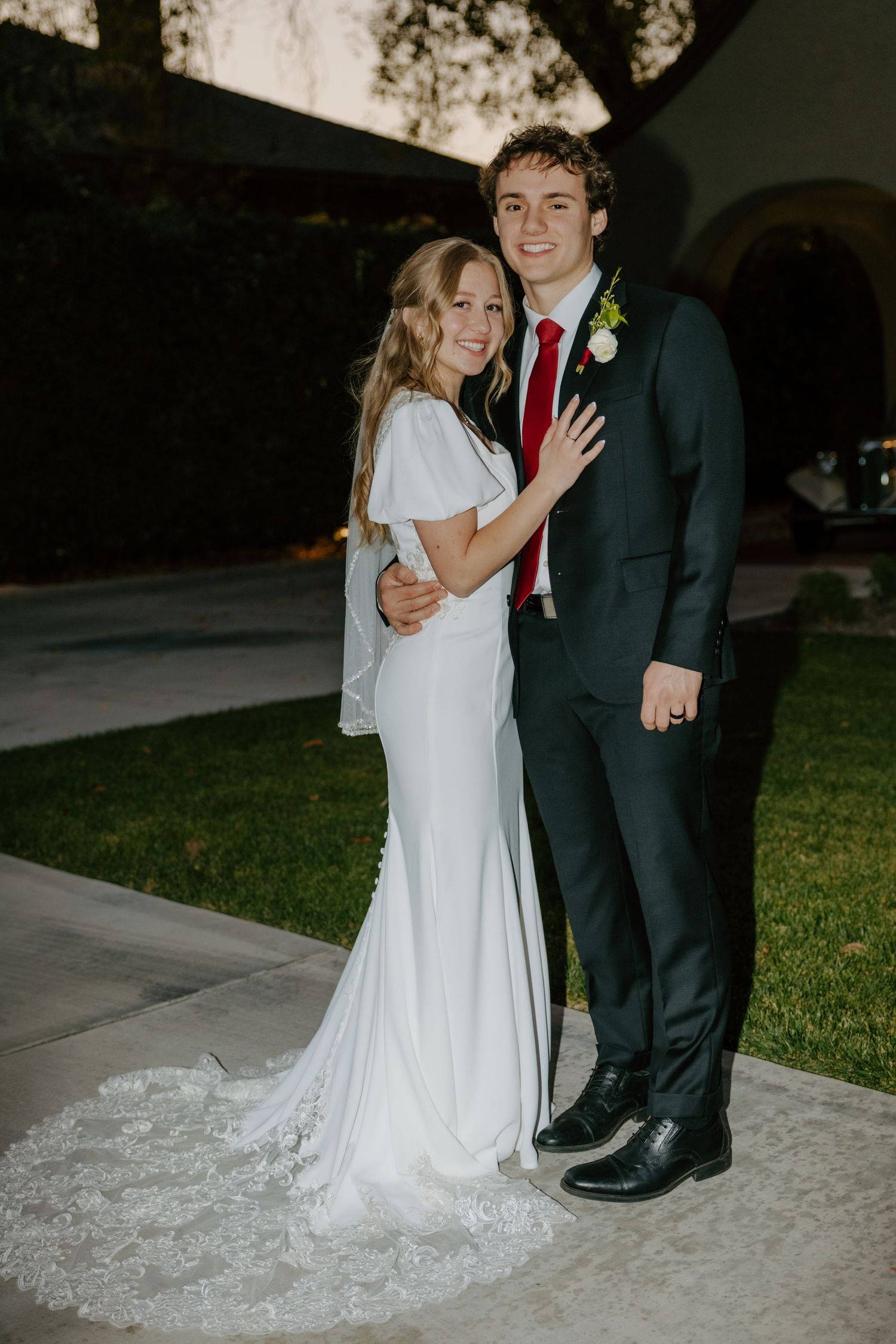 Bride in white lace gown and groom in dark suit with red tie pose together at outdoor wedding.