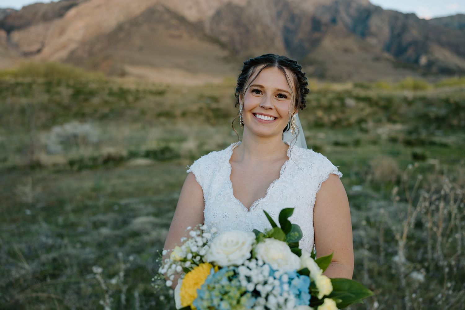 Smiling bride in white lace gown holding bouquet of yellow and blue flowers with mountain landscape background.