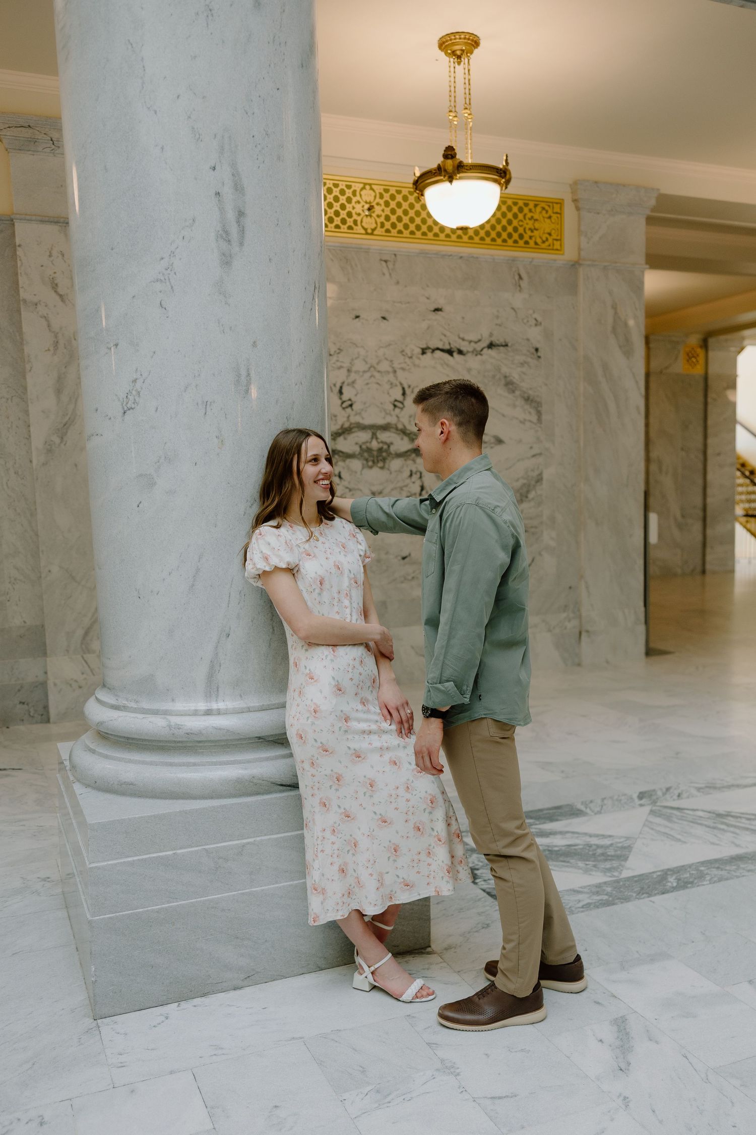 Man tenderly touching womans face as she leans against marble column in ornate building interior.
