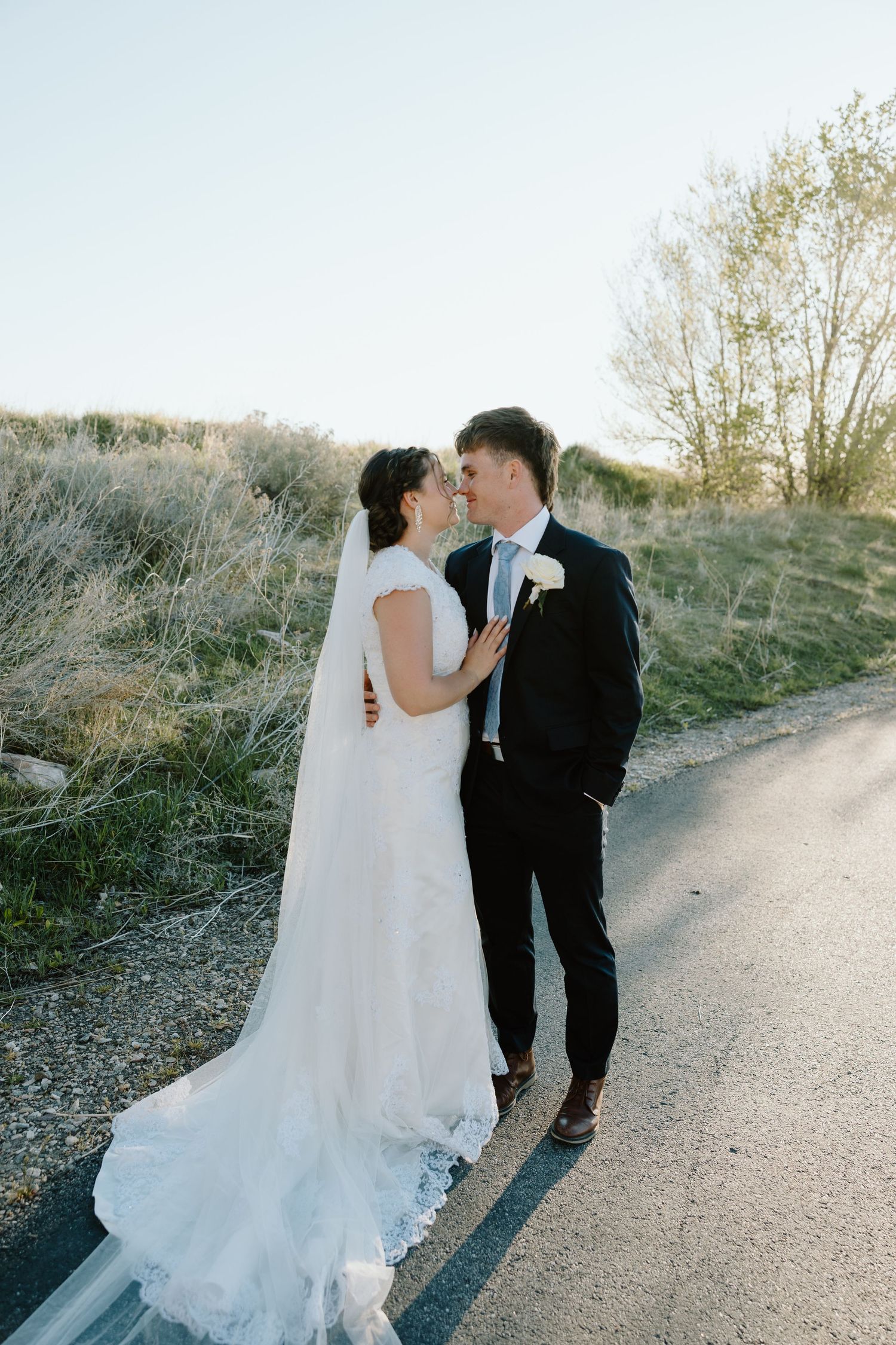 Bride and groom share a romantic moment on a country road, bride in elegant lace gown with veil.