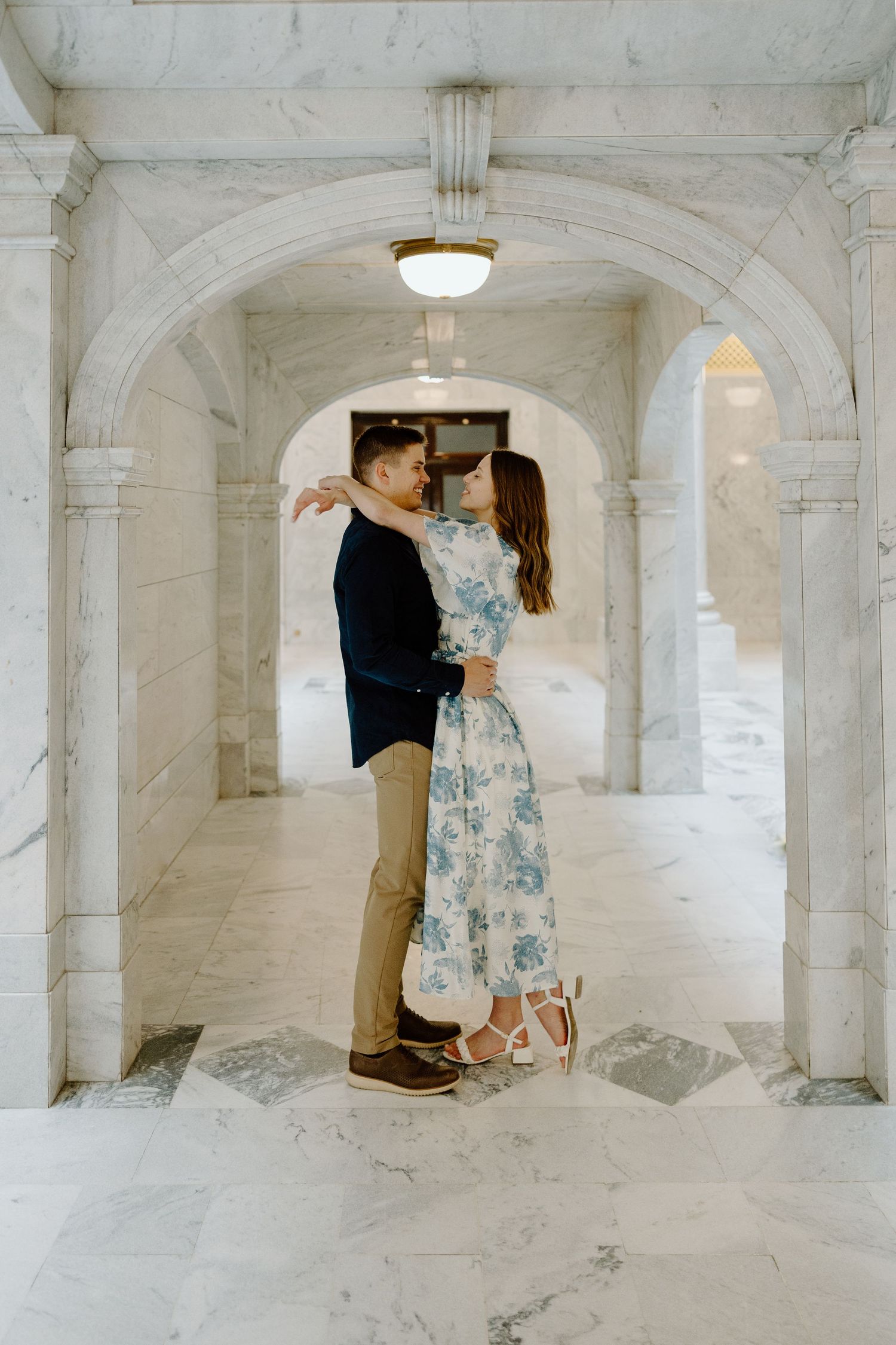 Couple embracing romantically in elegant marble archway corridor, woman in floral dress, man in navy blazer.