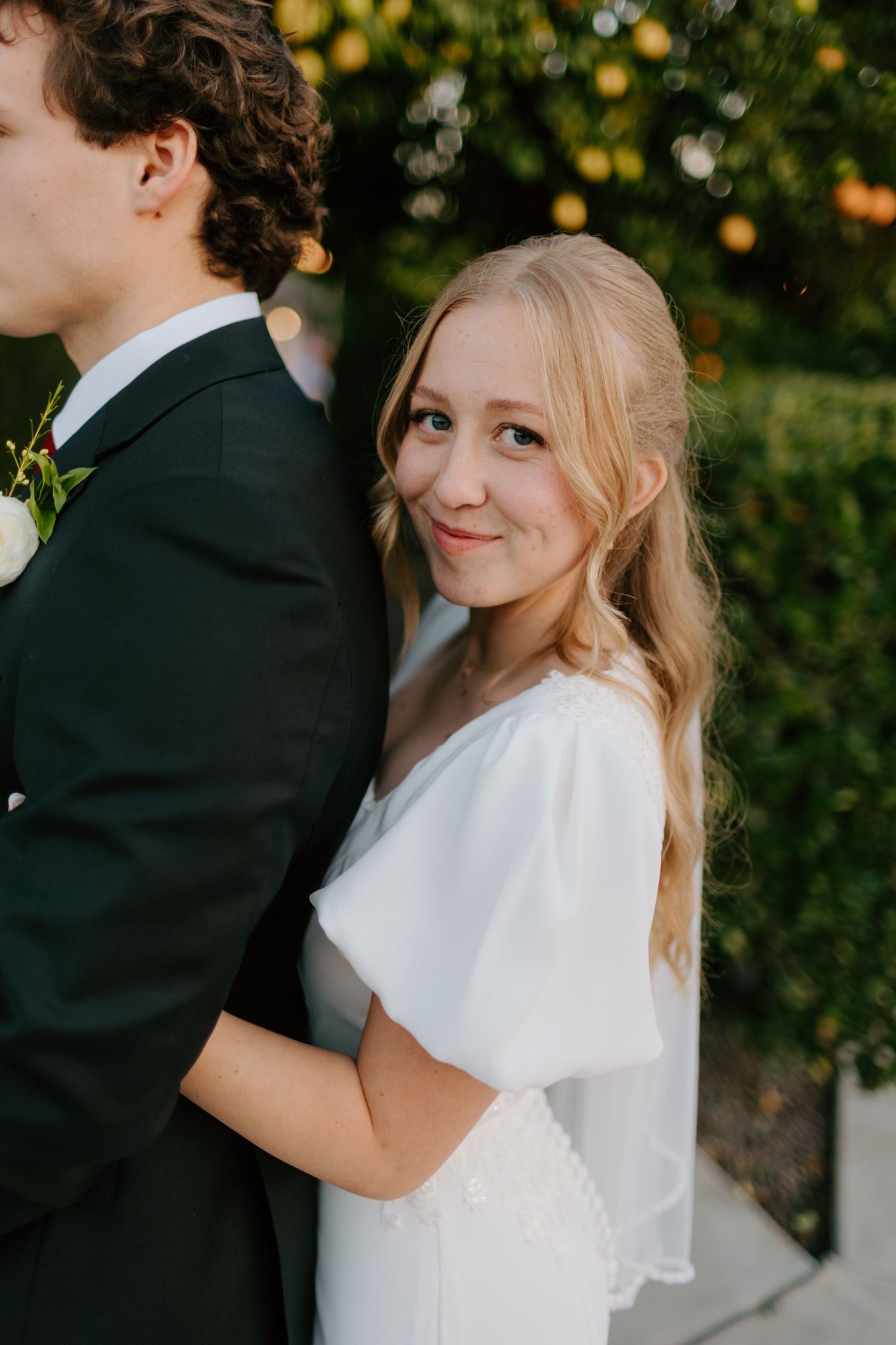 Smiling blonde bride in white flutter-sleeve wedding dress hugging groom in dark suit, outdoor garden setting with warm lighting.
