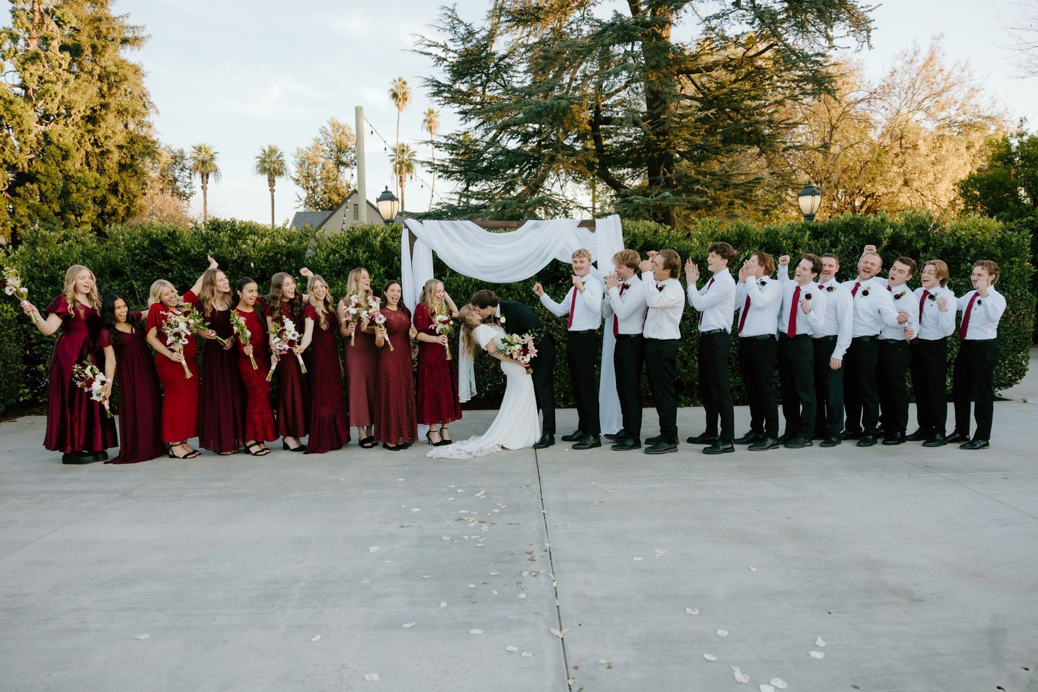 Large wedding party poses with bride and groom, bridesmaids in burgundy and groomsmen in white shirts outdoors.