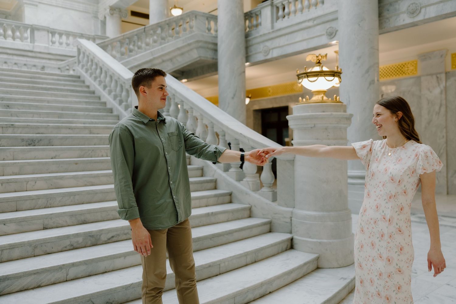 Couple holding hands on grand marble staircase in elegant building, man in green shirt, woman in floral dress.