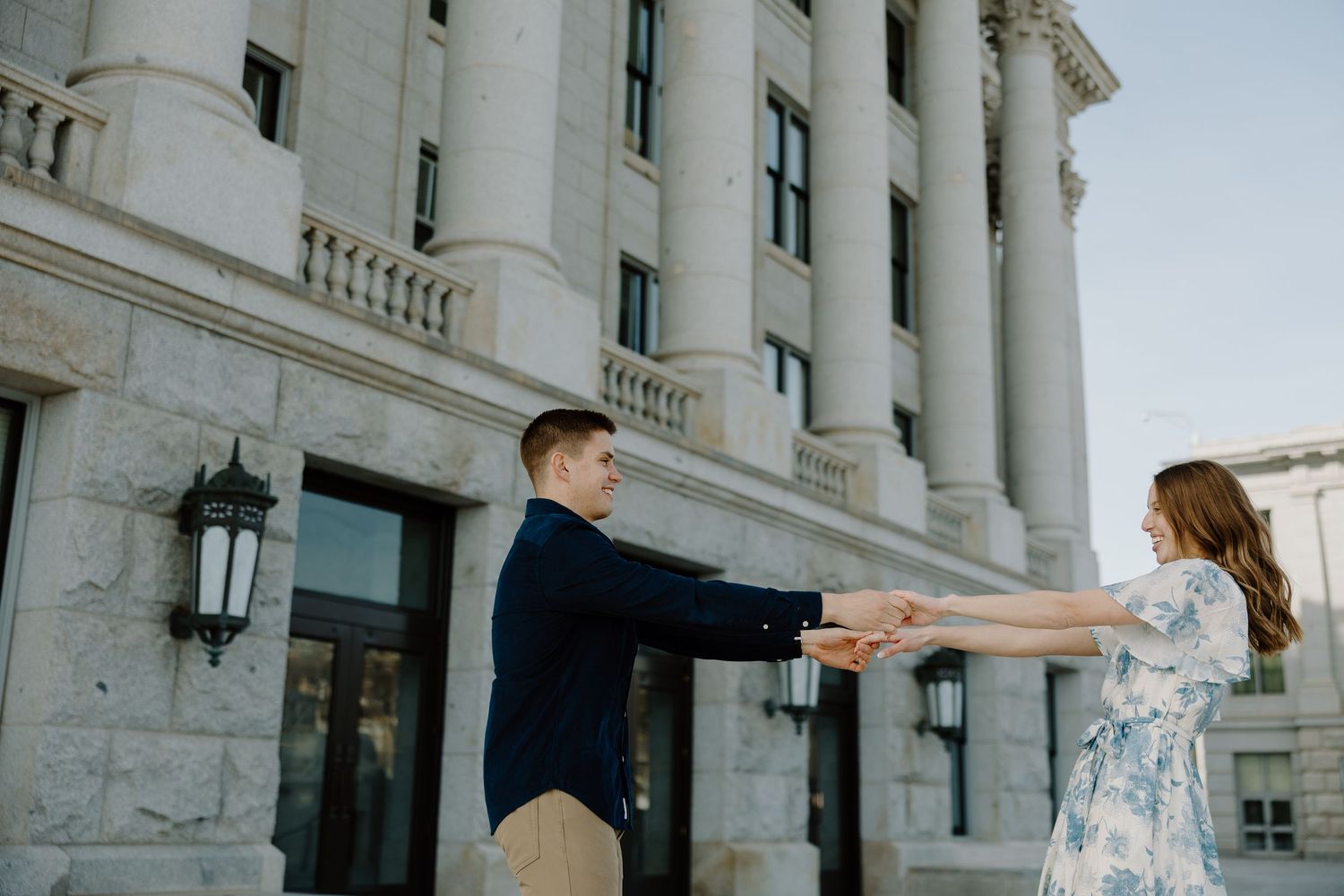 Engaged couple holding hands playfully outside grand stone columned building in golden hour light.