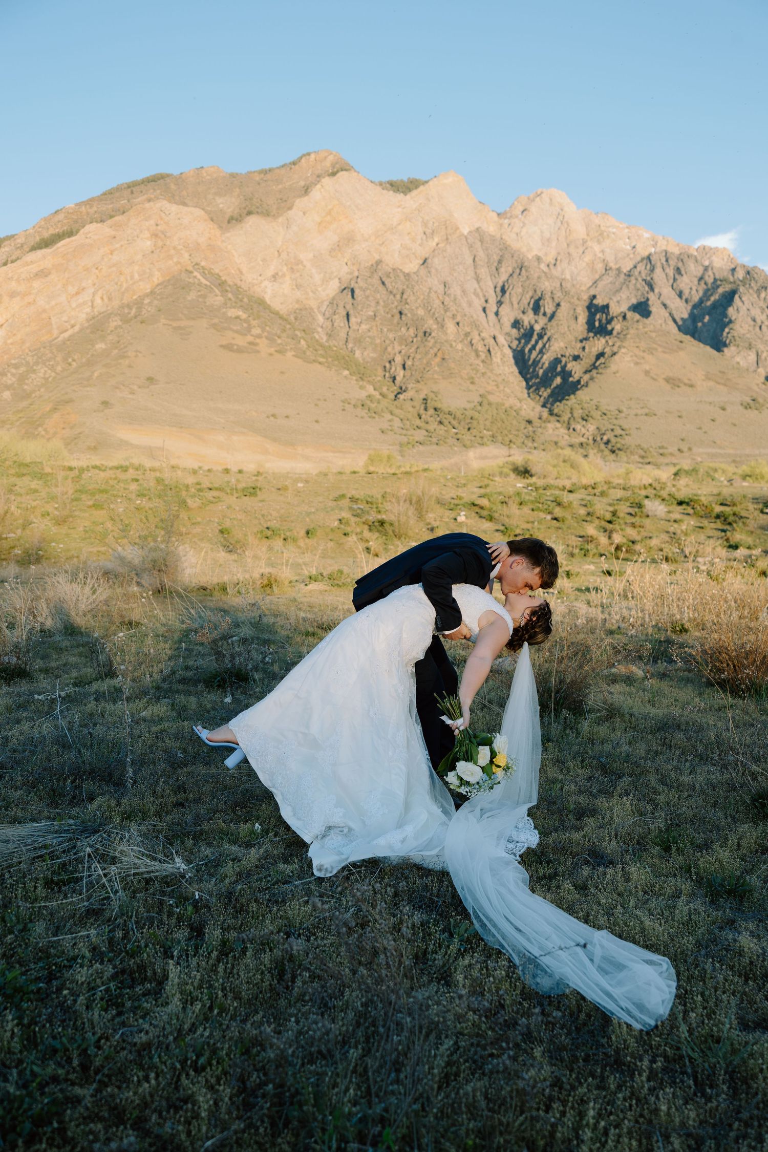 Groom dipping bride for a romantic kiss in an open field with dramatic mountain backdrop at sunset.