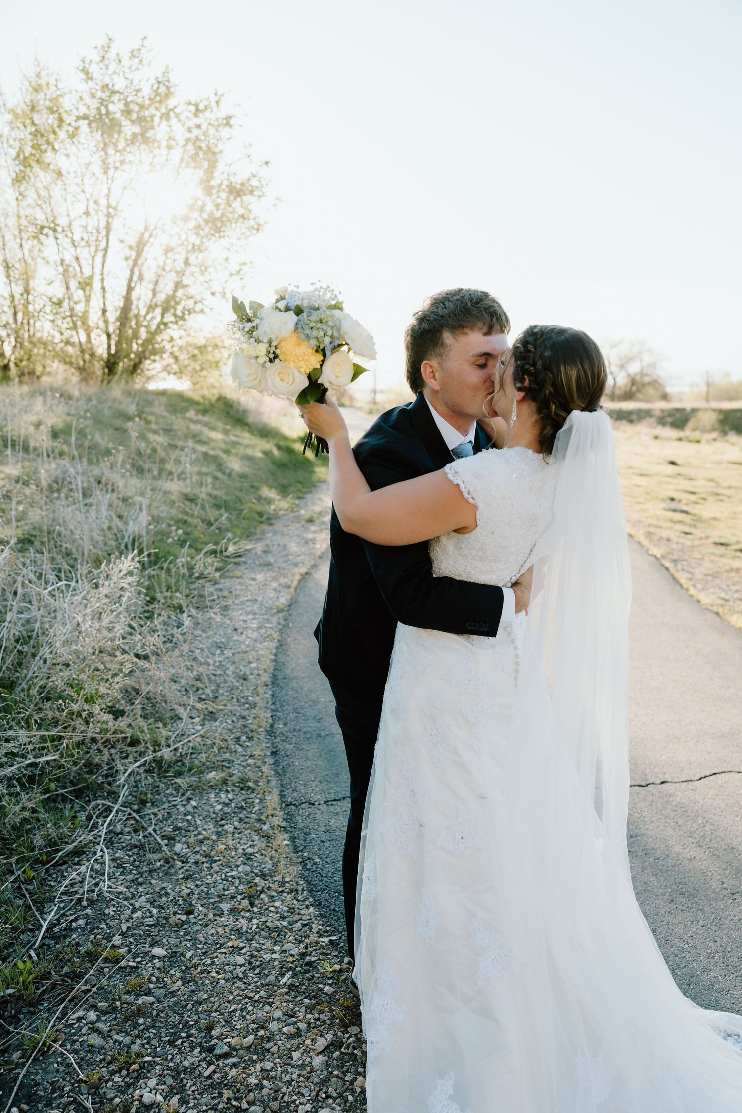 Bride and groom sharing romantic kiss on pathway, bride holding white flower bouquet.