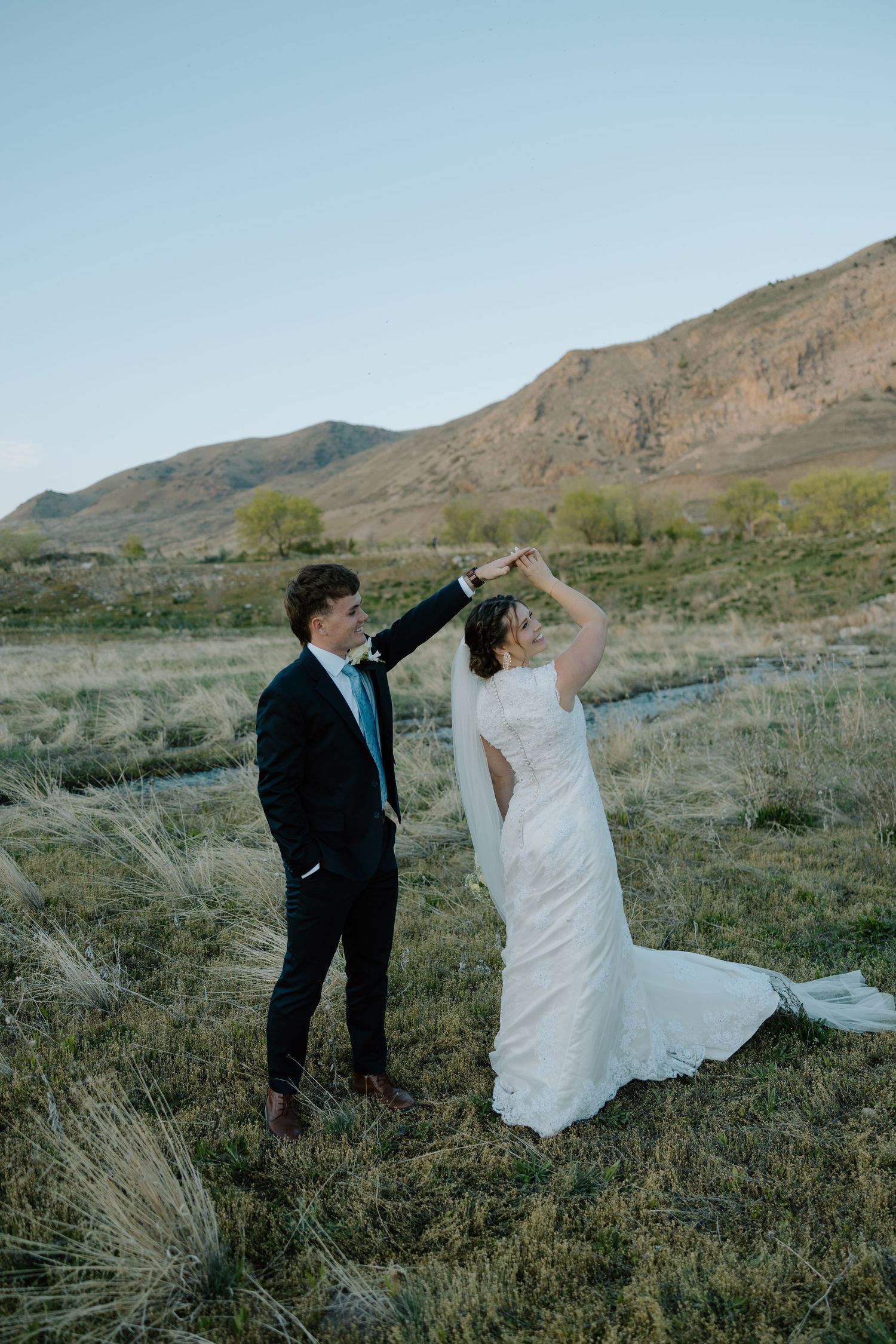 Bride and groom dancing outdoors in a scenic mountain field, bride in white gown with veil, groom in dark suit.