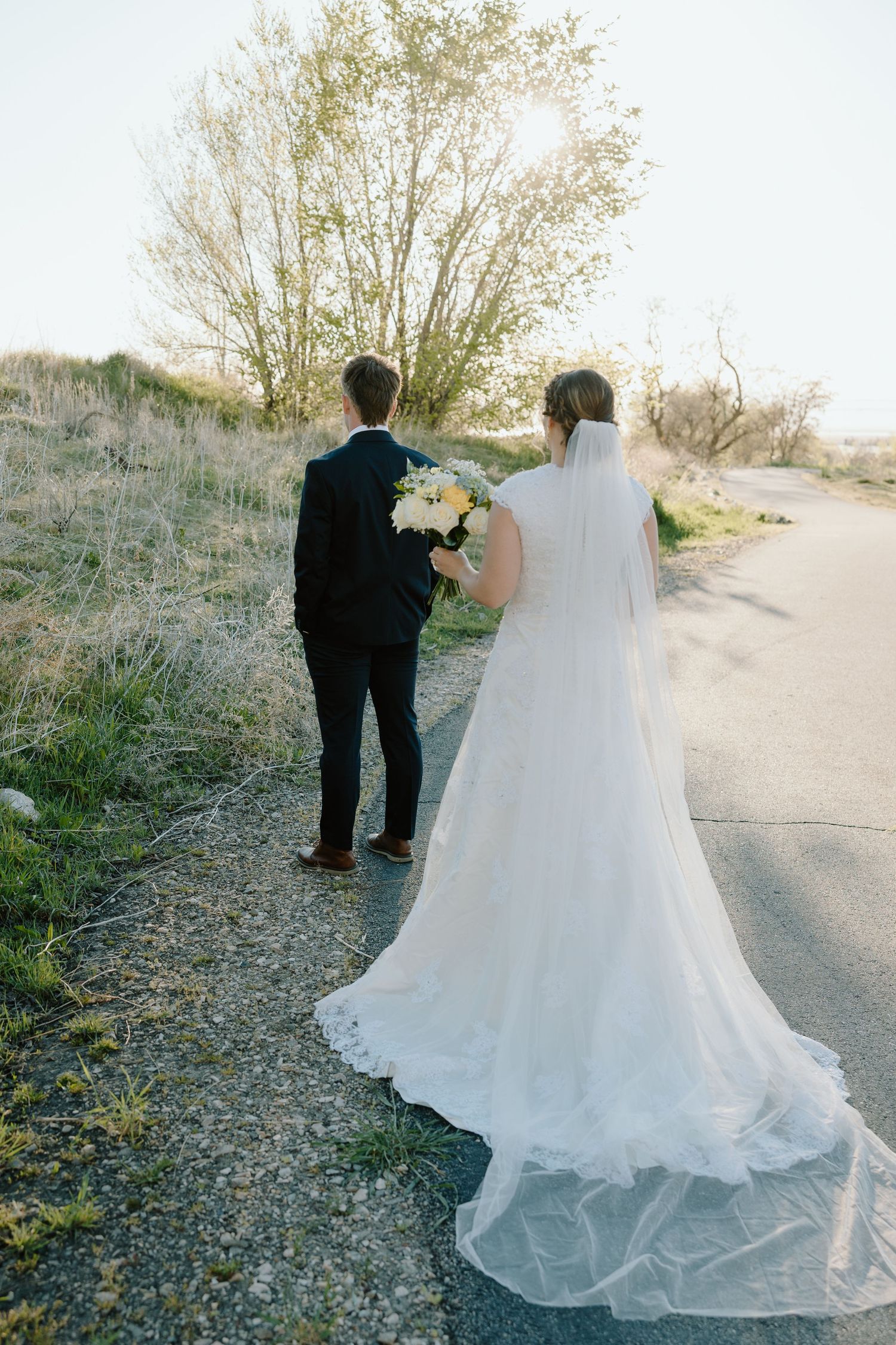 Bride and groom walking hand in hand down a country road, bride wearing a flowing white gown with long veil.