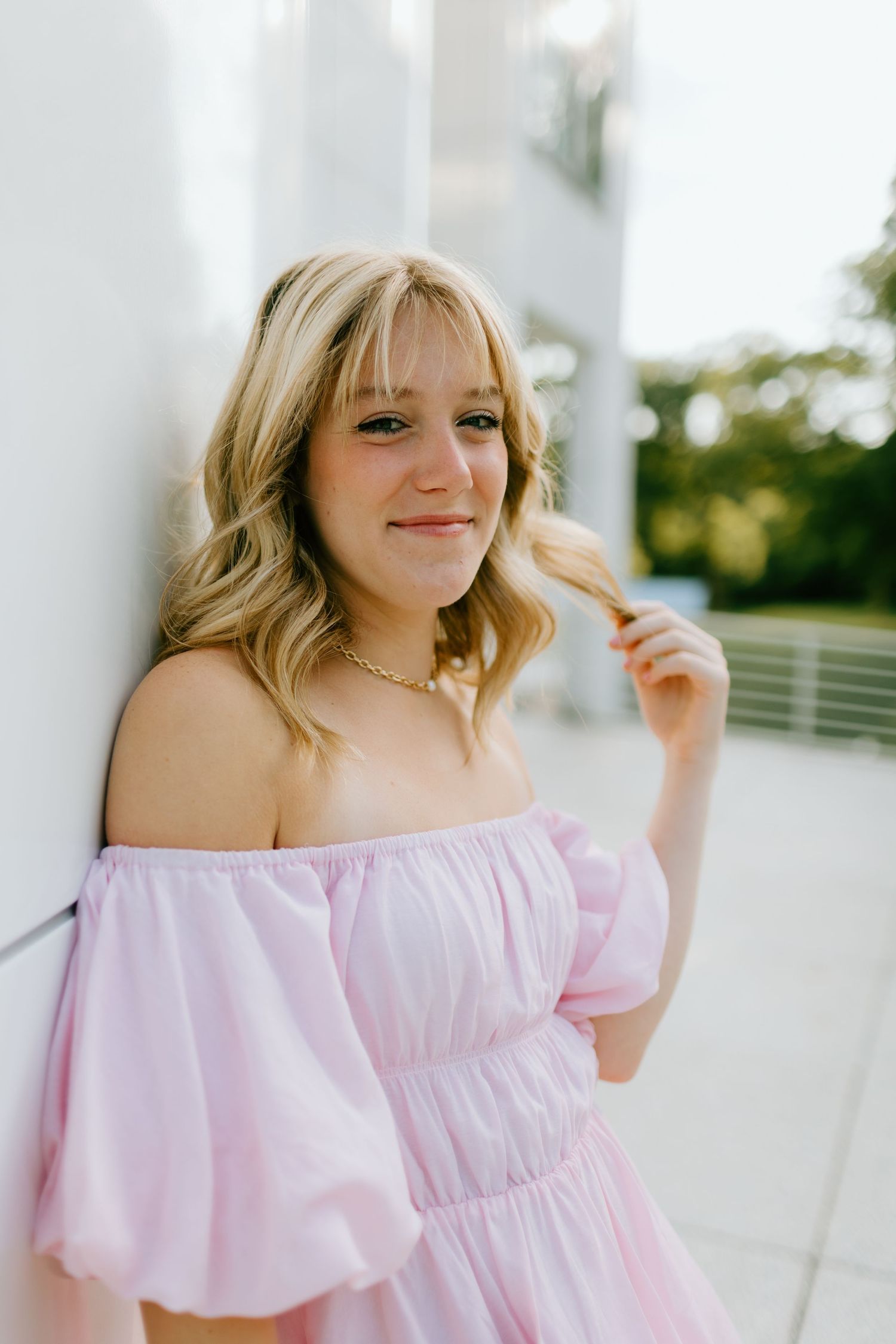 Young blonde woman in pink off-shoulder dress poses outdoors, smiling softly while touching her hair.