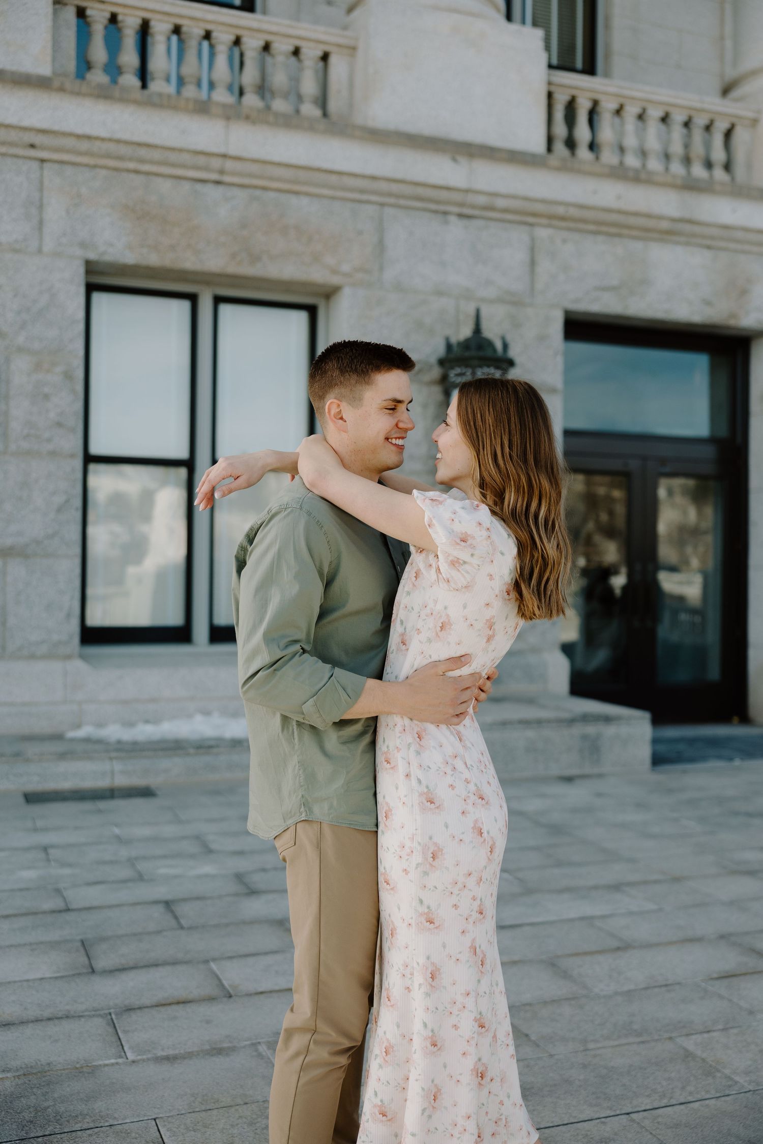 A couple embraces and smiles at each other outside a grand stone building during an engagement photo session.