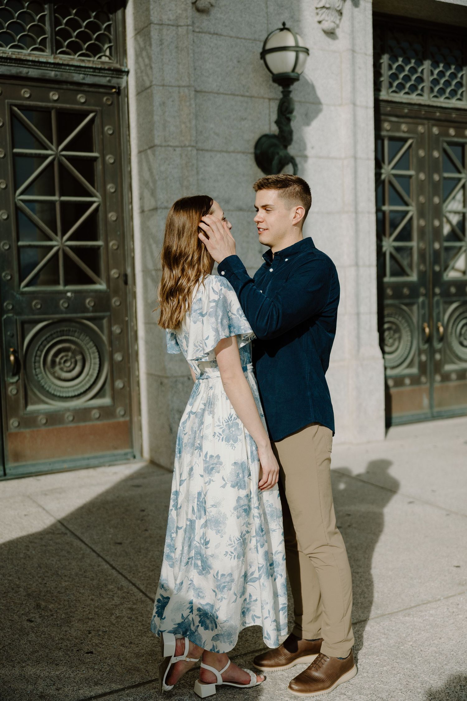 Man gently brushing woman's hair back outside ornate bronze doors of a classical stone building.