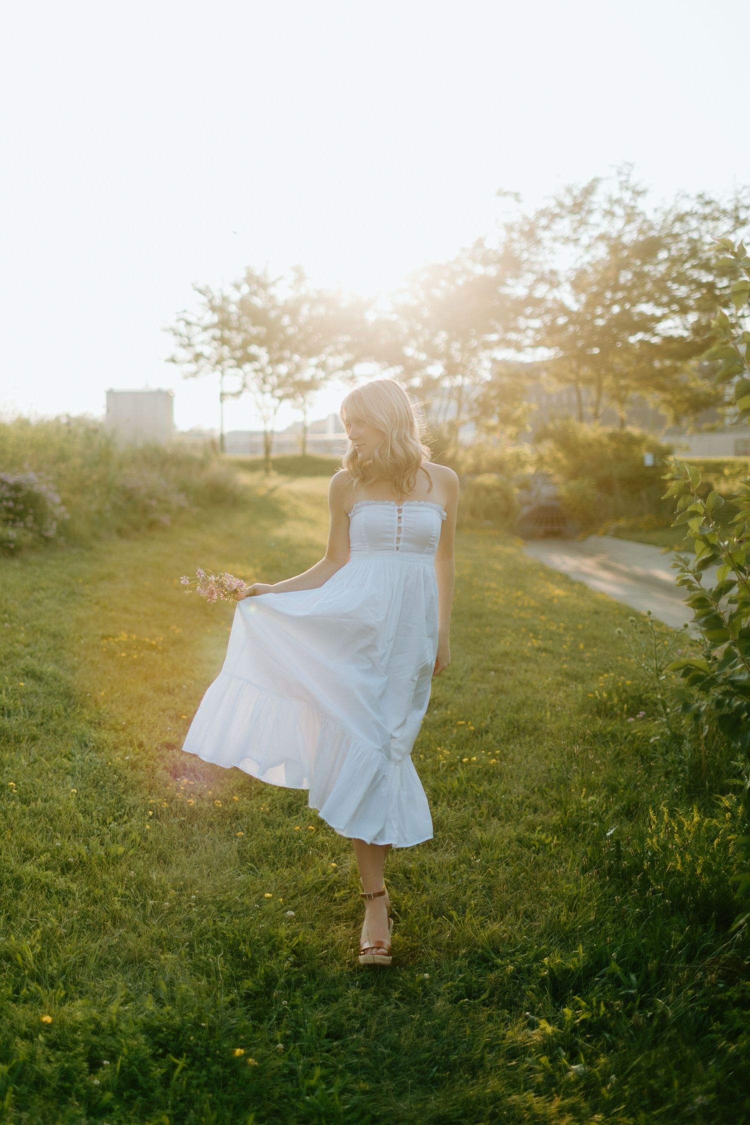 Woman in white dress holding flowers, twirling in sunlit green field at golden hour.