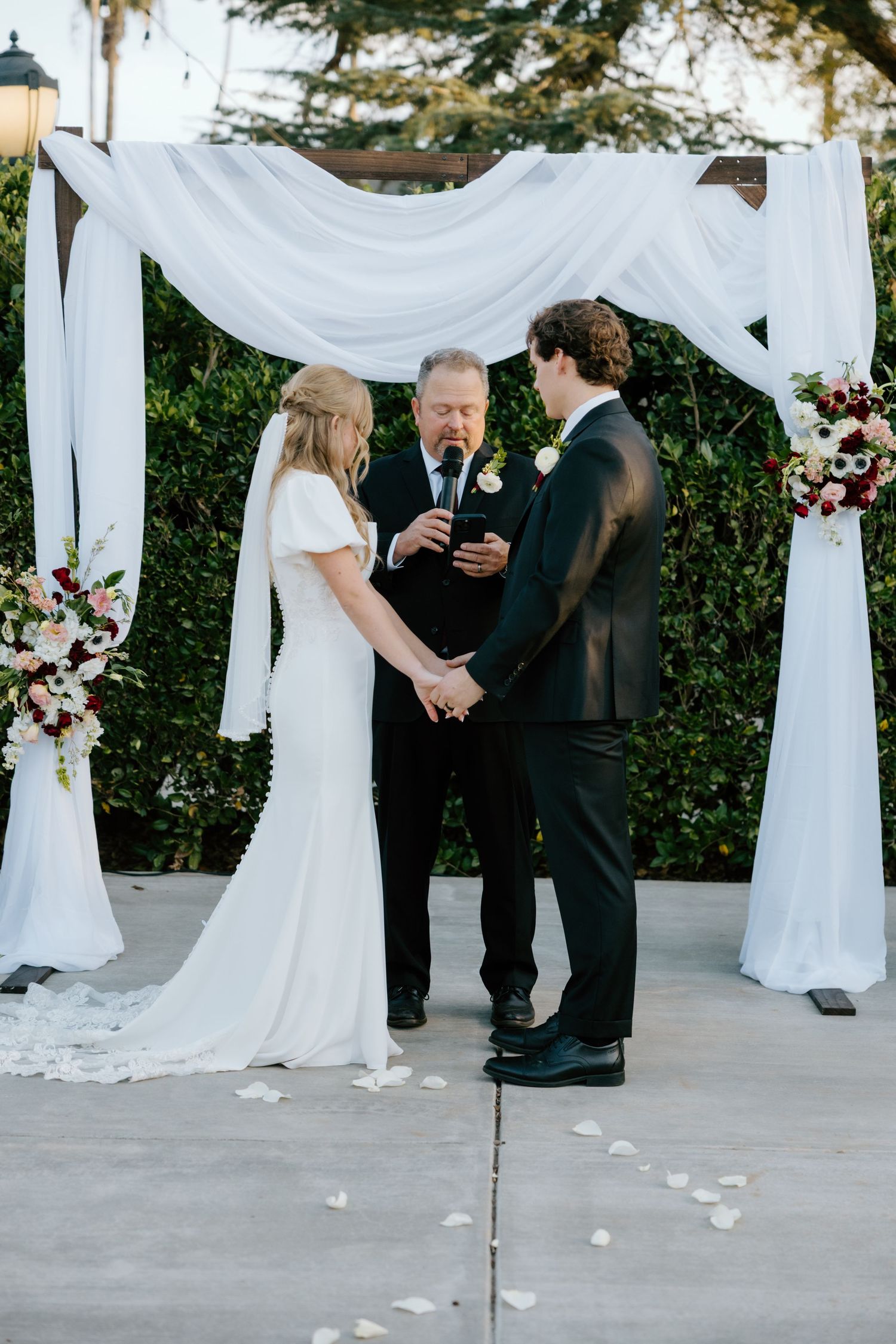 Bride and groom hold hands as officiant reads vows under a white draped arch at outdoor wedding ceremony.