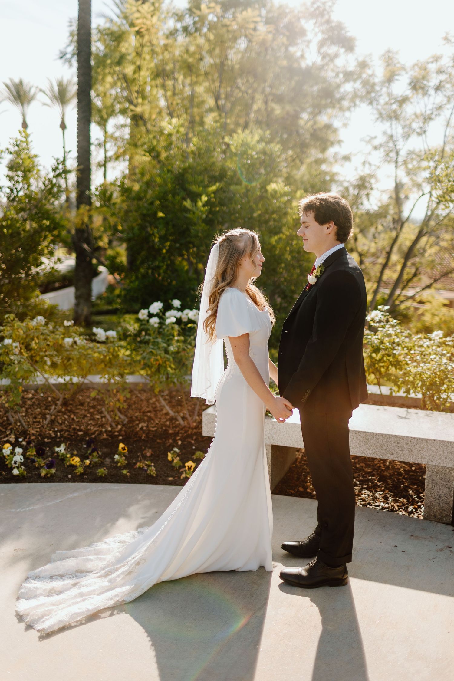 Bride and groom hold hands in sunlit garden, she wears white gown with veil, he wears black tuxedo.