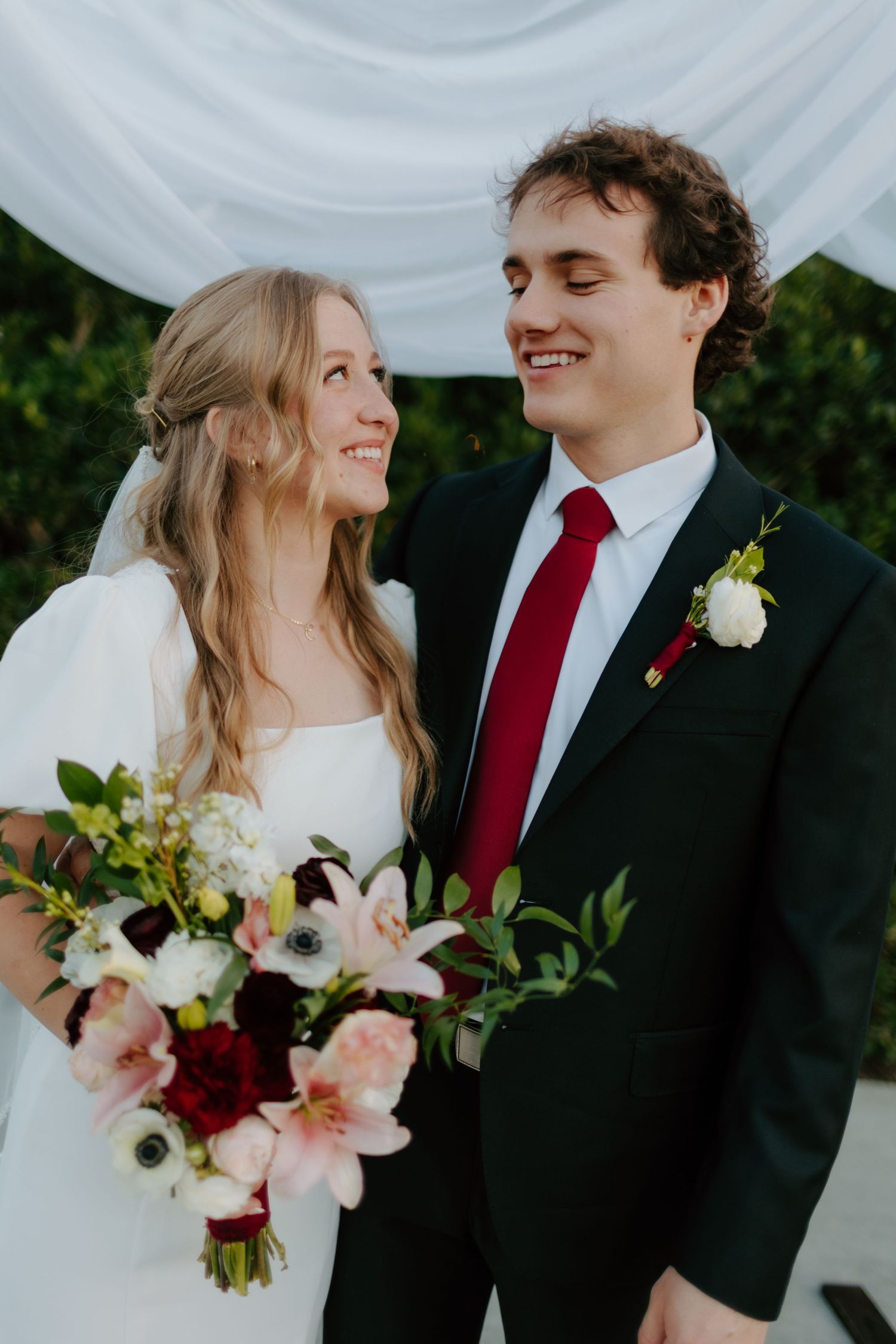 Smiling bride and groom in black suit with red tie sharing a tender moment at outdoor wedding ceremony.