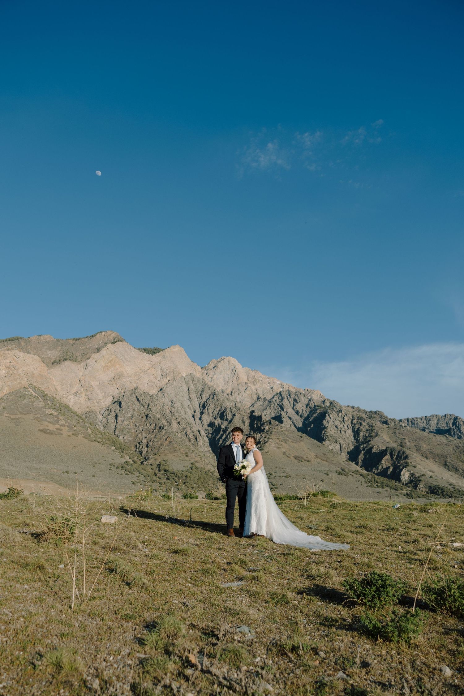Bride and groom posing together in mountain landscape under clear blue sky with moon.