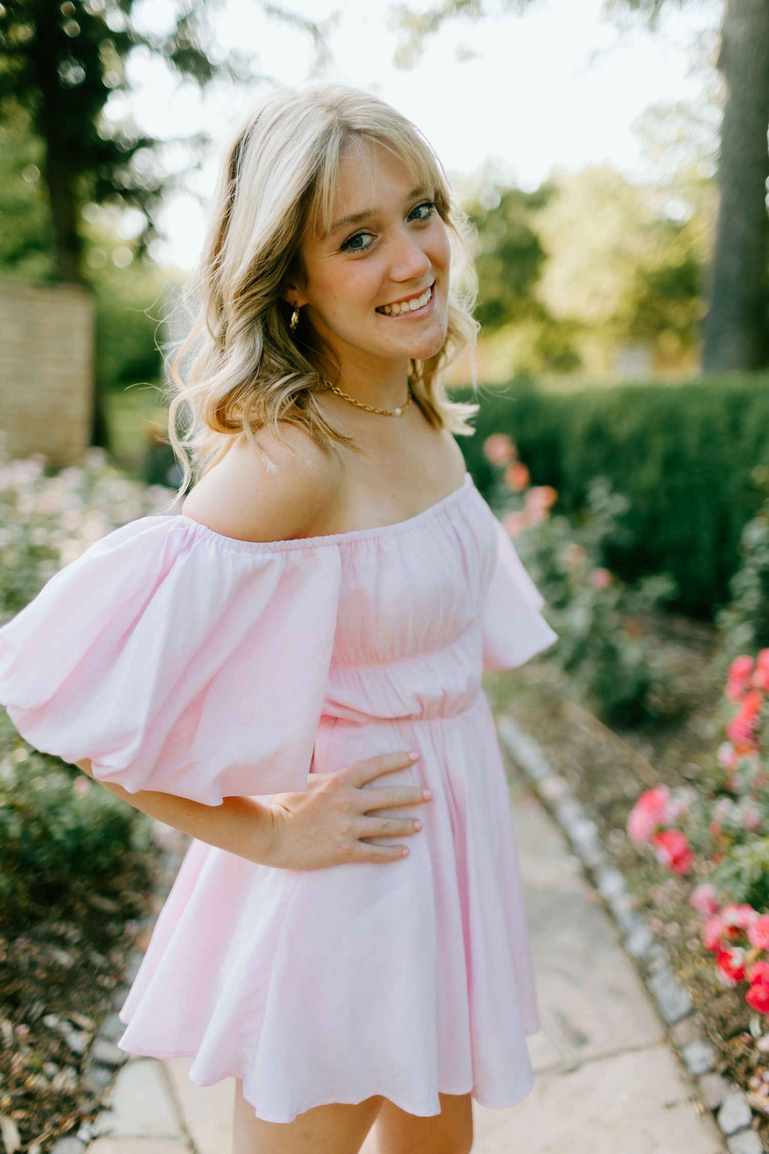 Young woman in pink off-shoulder dress smiling in a garden with roses.