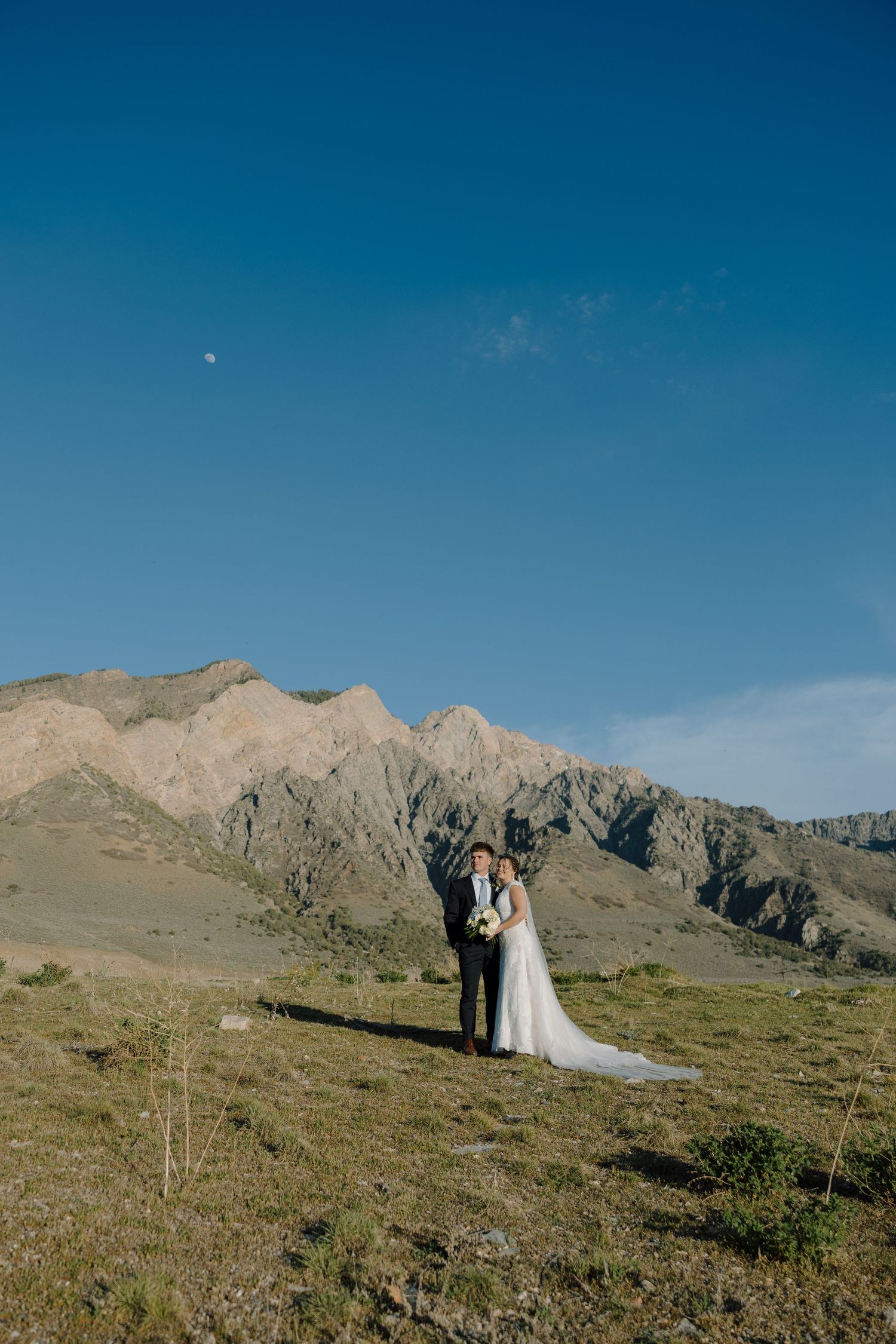 Wedding couple standing in vast mountain meadow with dramatic rocky peaks behind them.